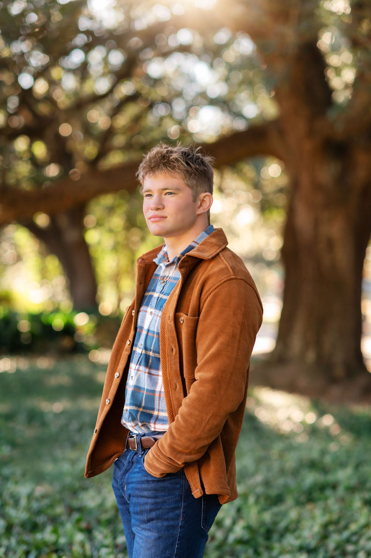 Young man in brown suede jacket and plaid shirt standing outdoors among trees in a senior portrait photo.