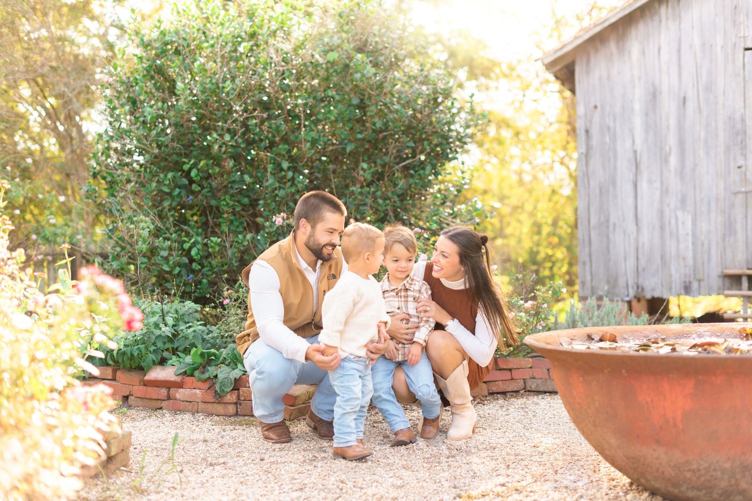 Family of four sitting together in a garden patio setting, parents and two young children smiling warmly.