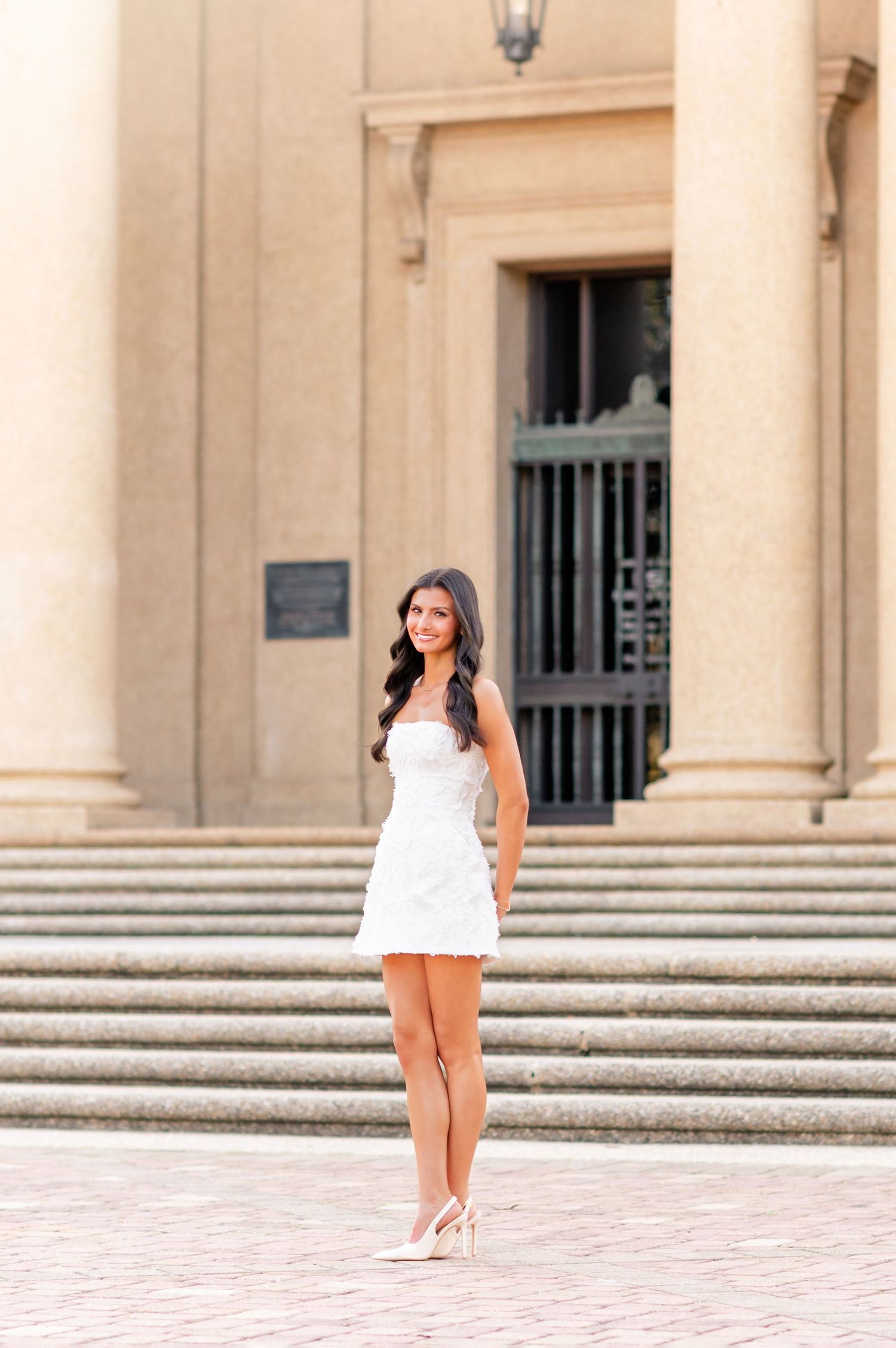 Young woman in white mini dress and heels poses on plaza steps of classical stone building.
