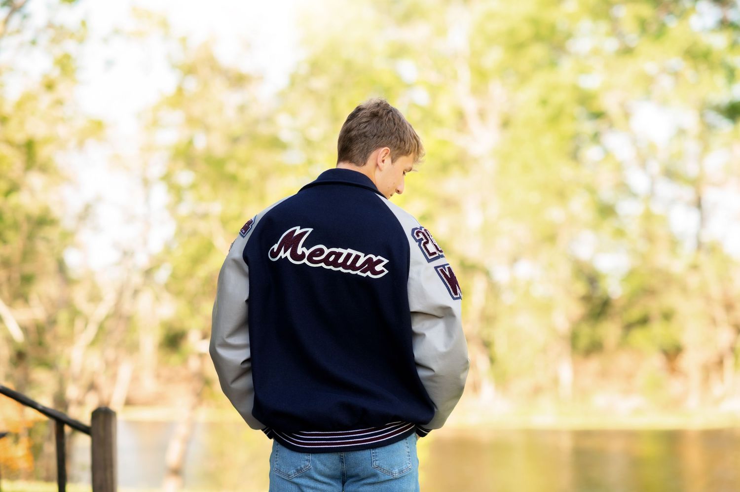 Teen boy wearing a navy and gray Meaux varsity letterman jacket, standing outdoors near a pond.
