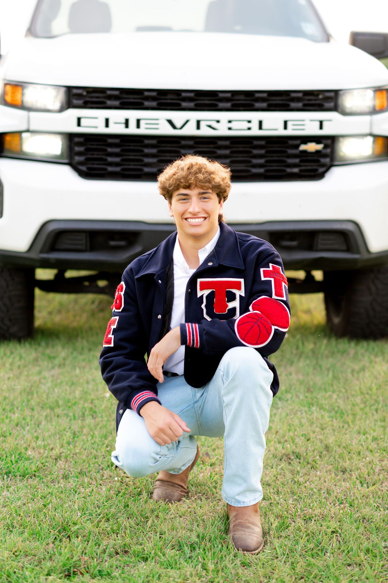 Smiling teen in letterman jacket crouches in front of white Chevrolet truck on grass for senior photos.
