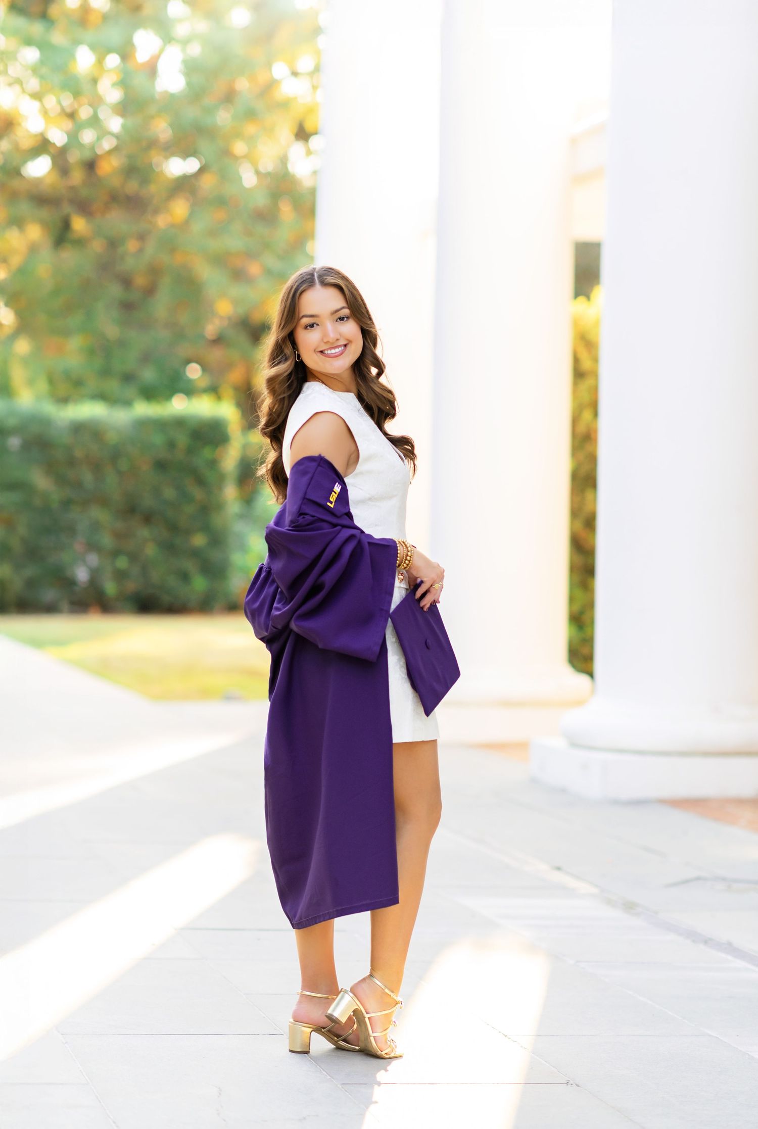 Young woman in white dress holding purple graduation gown, posing between white columns outdoors.