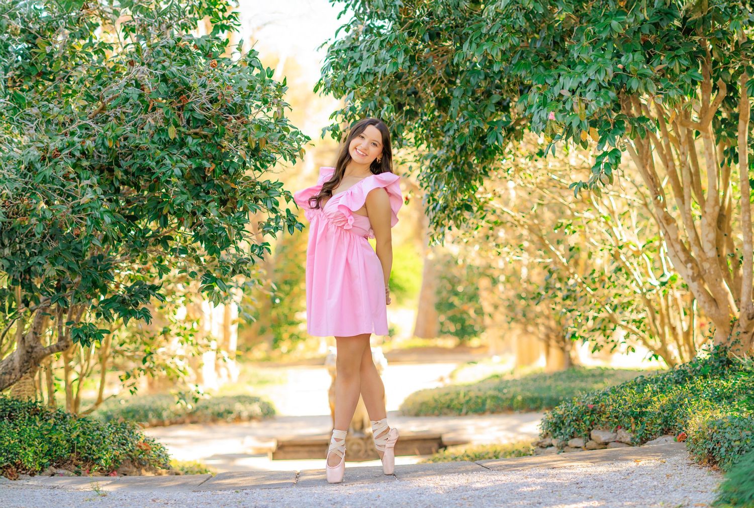 Young woman in pink dress poses en pointe on a garden path surrounded by lush green trees.