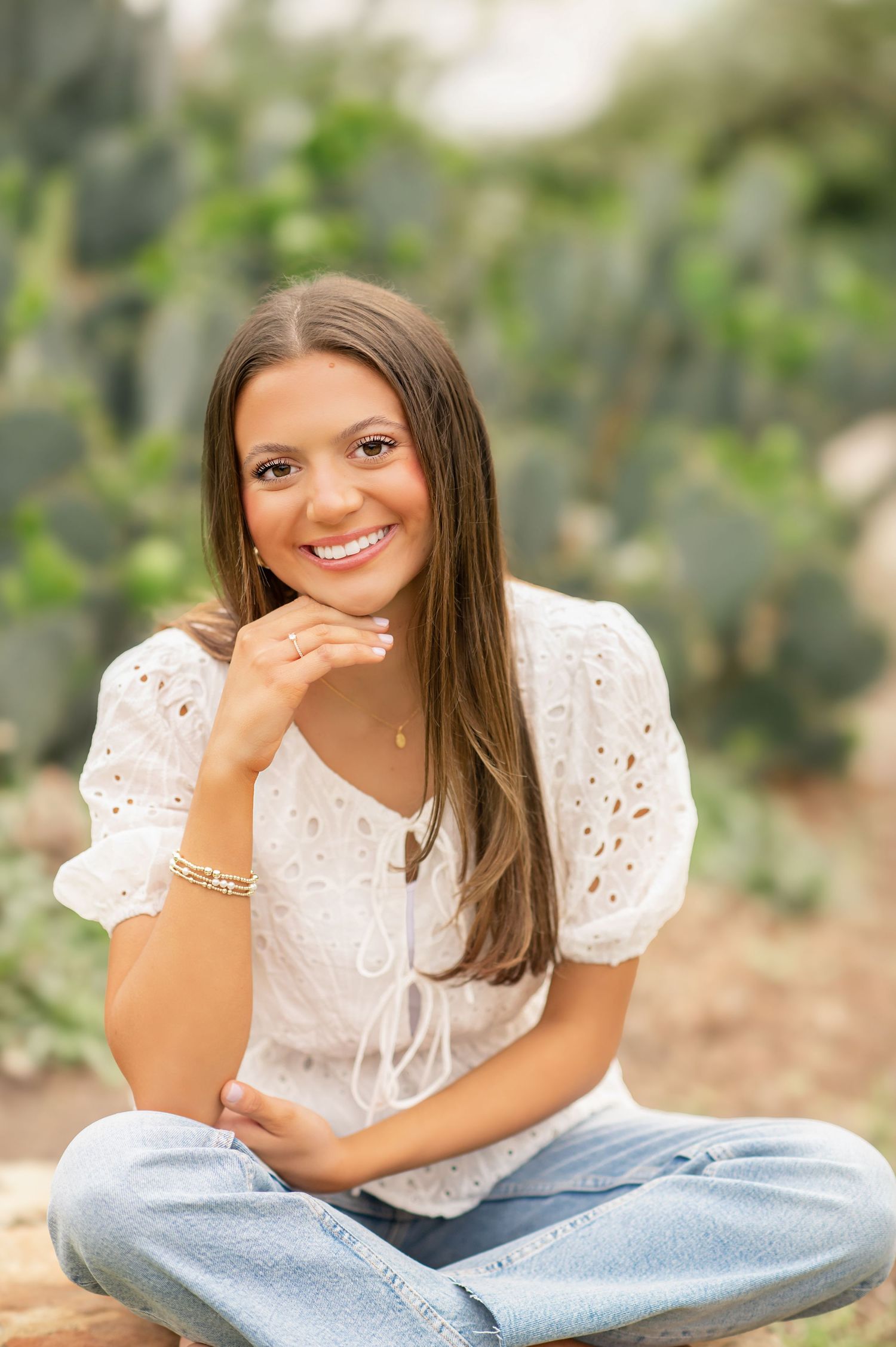Brunette girl in white eyelet top and jeans sitting cross-legged and smiling near cactus garden.