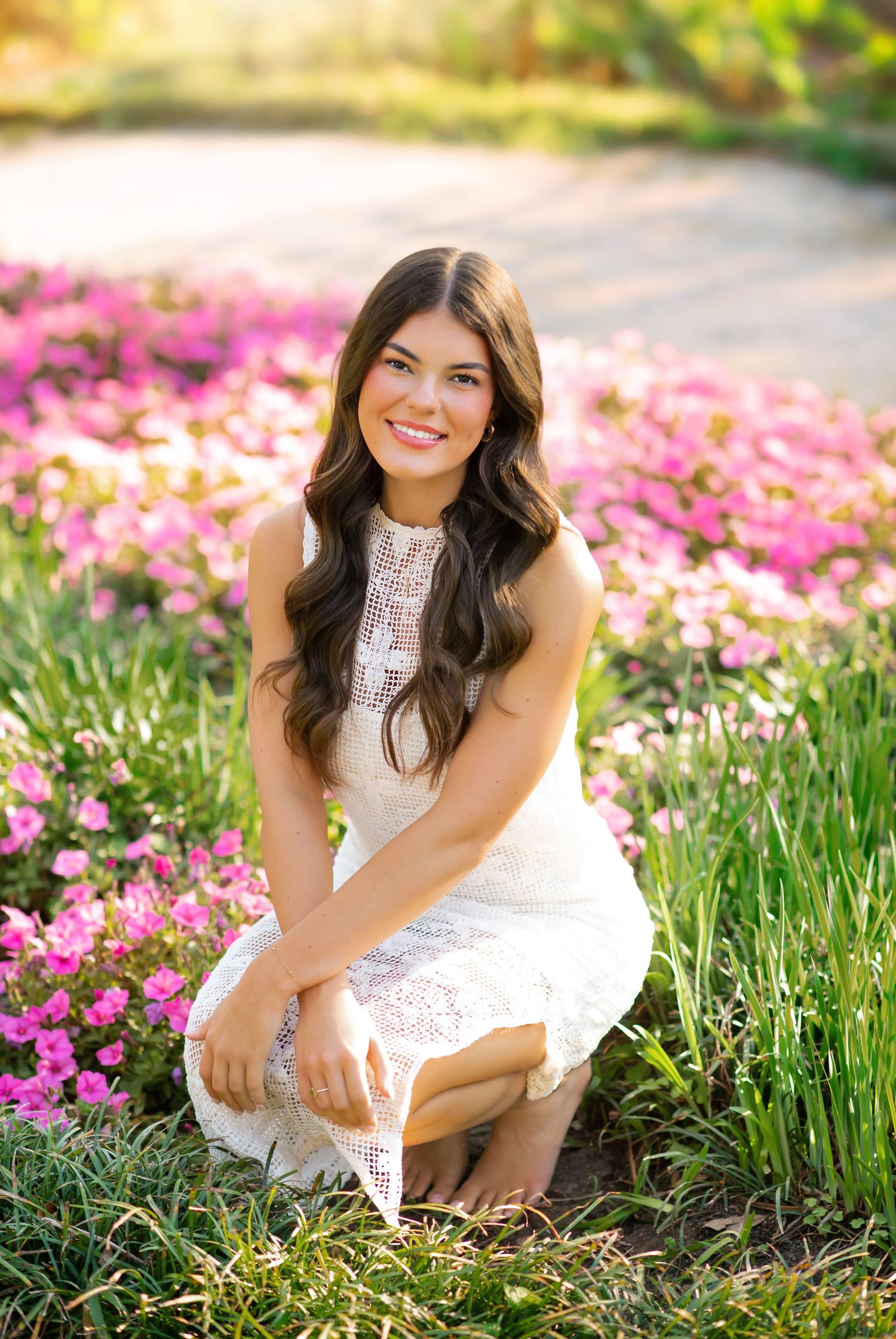 Young woman in white lace dress smiling among pink flowers in a sunny garden setting.