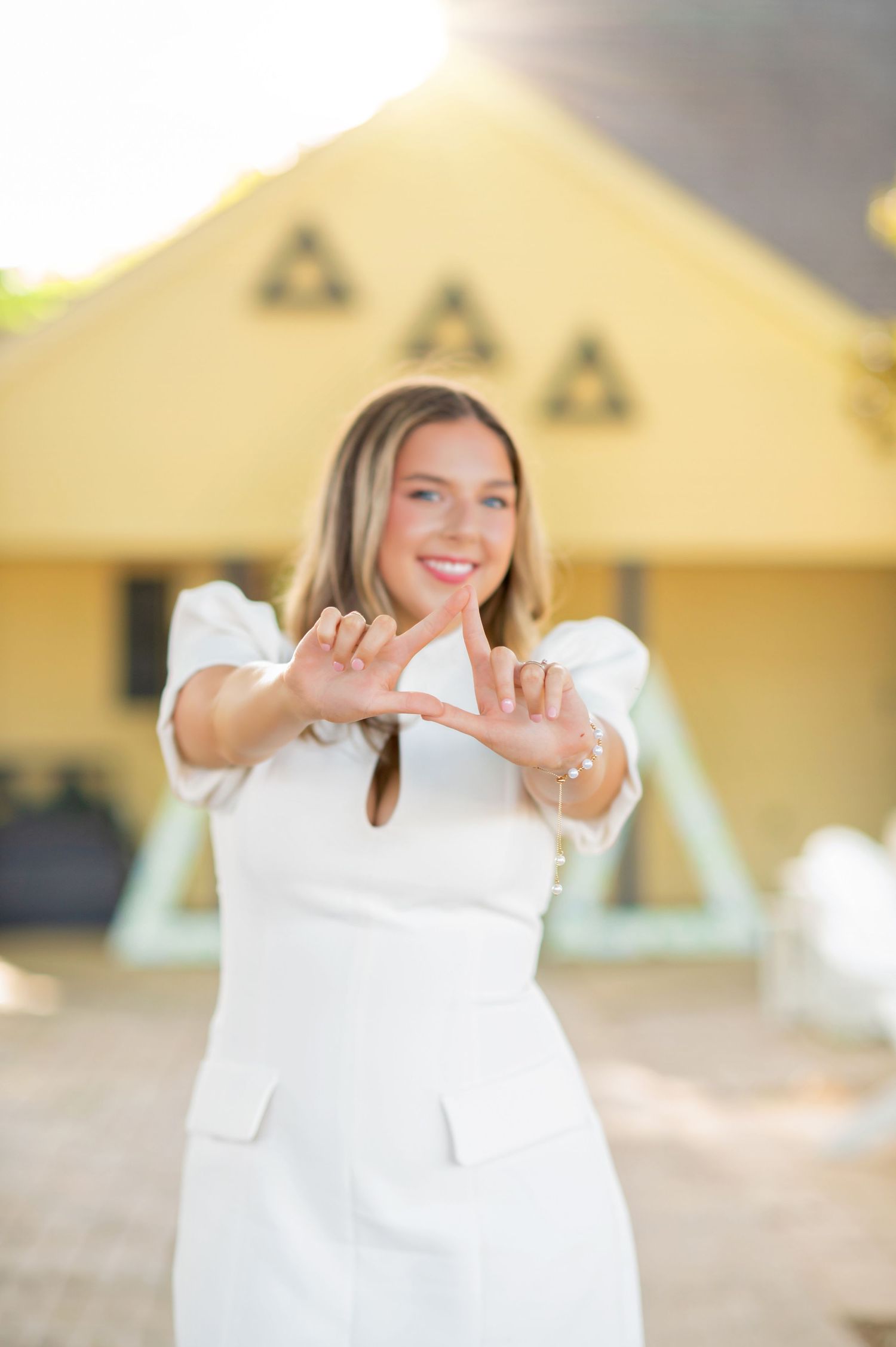 Smiling woman in white dress making triangle hand gesture in front of Greek letter sorority house.