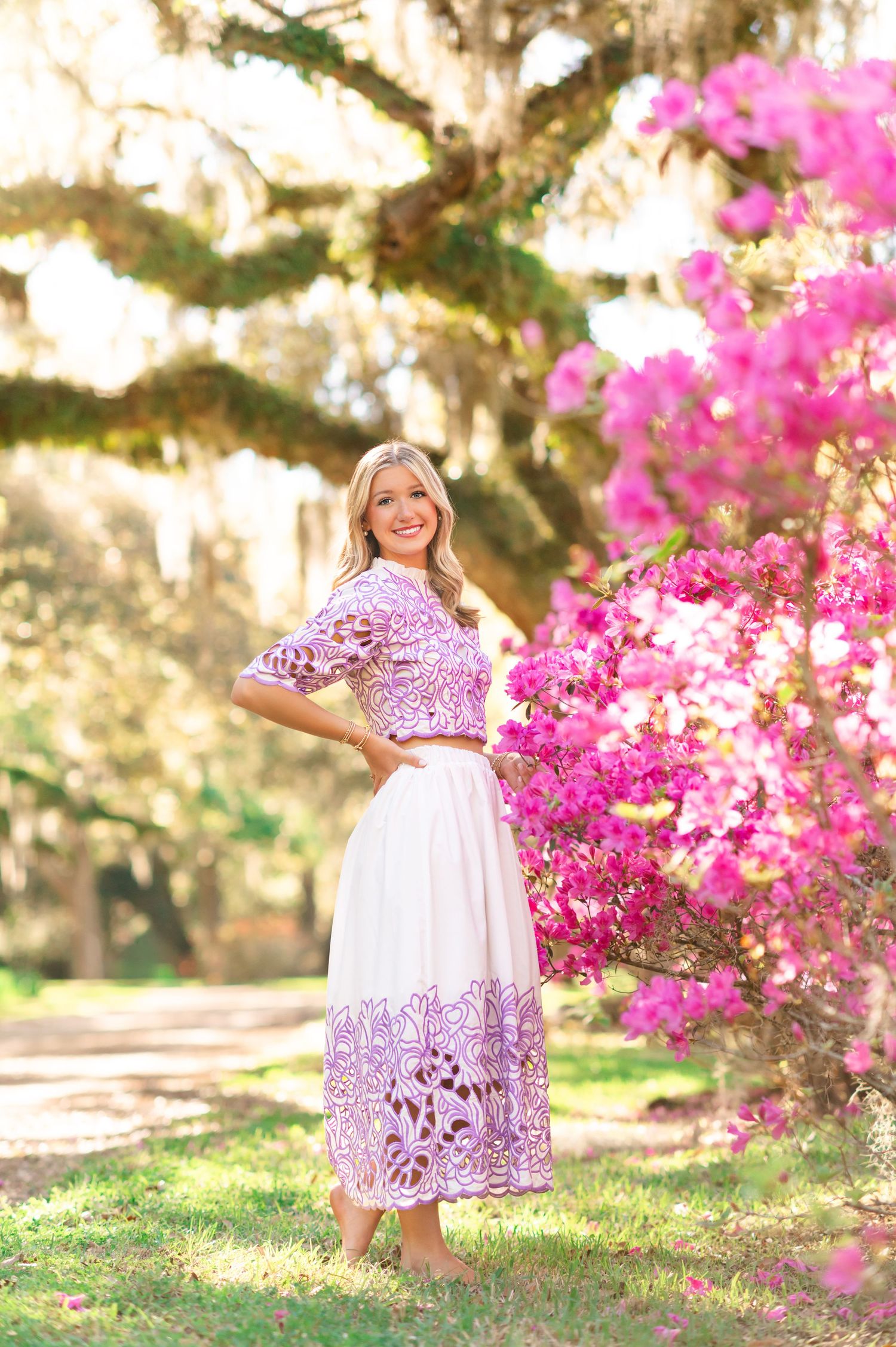 Young woman in purple and white embroidered dress poses near blooming pink azaleas under a moss-draped oak tree.