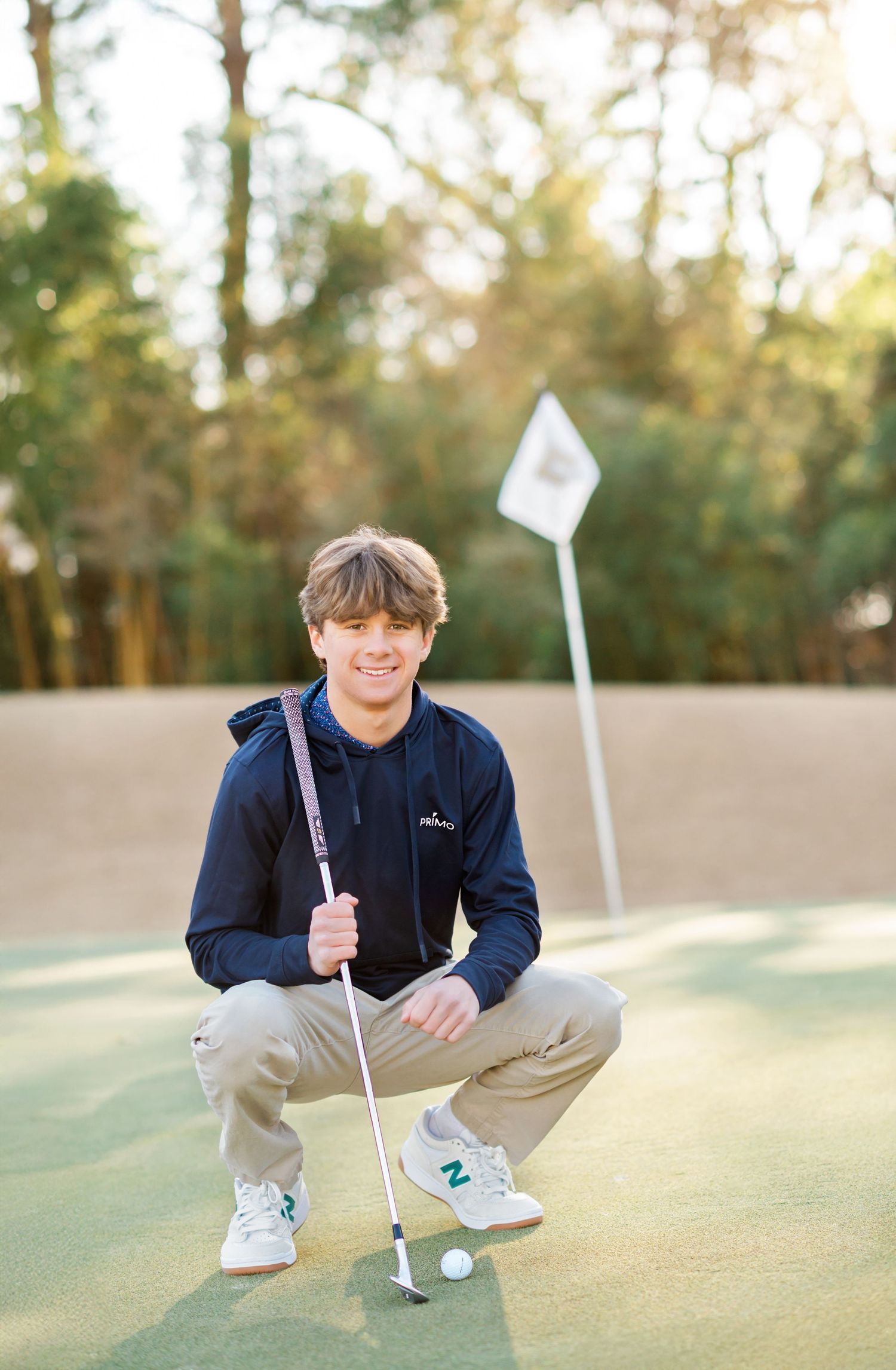 Young golfer crouching on putting green holding putter near golf ball with flag in background.
