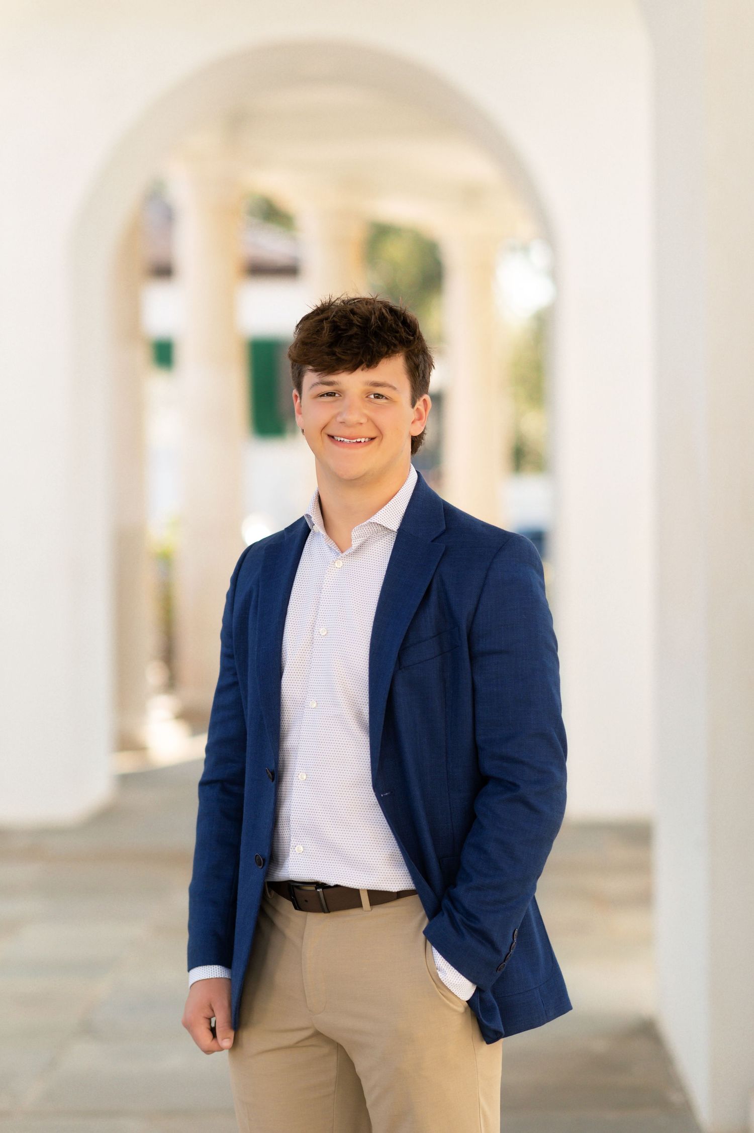 Young man in navy blazer and khakis smiling confidently under white arched colonnade.
