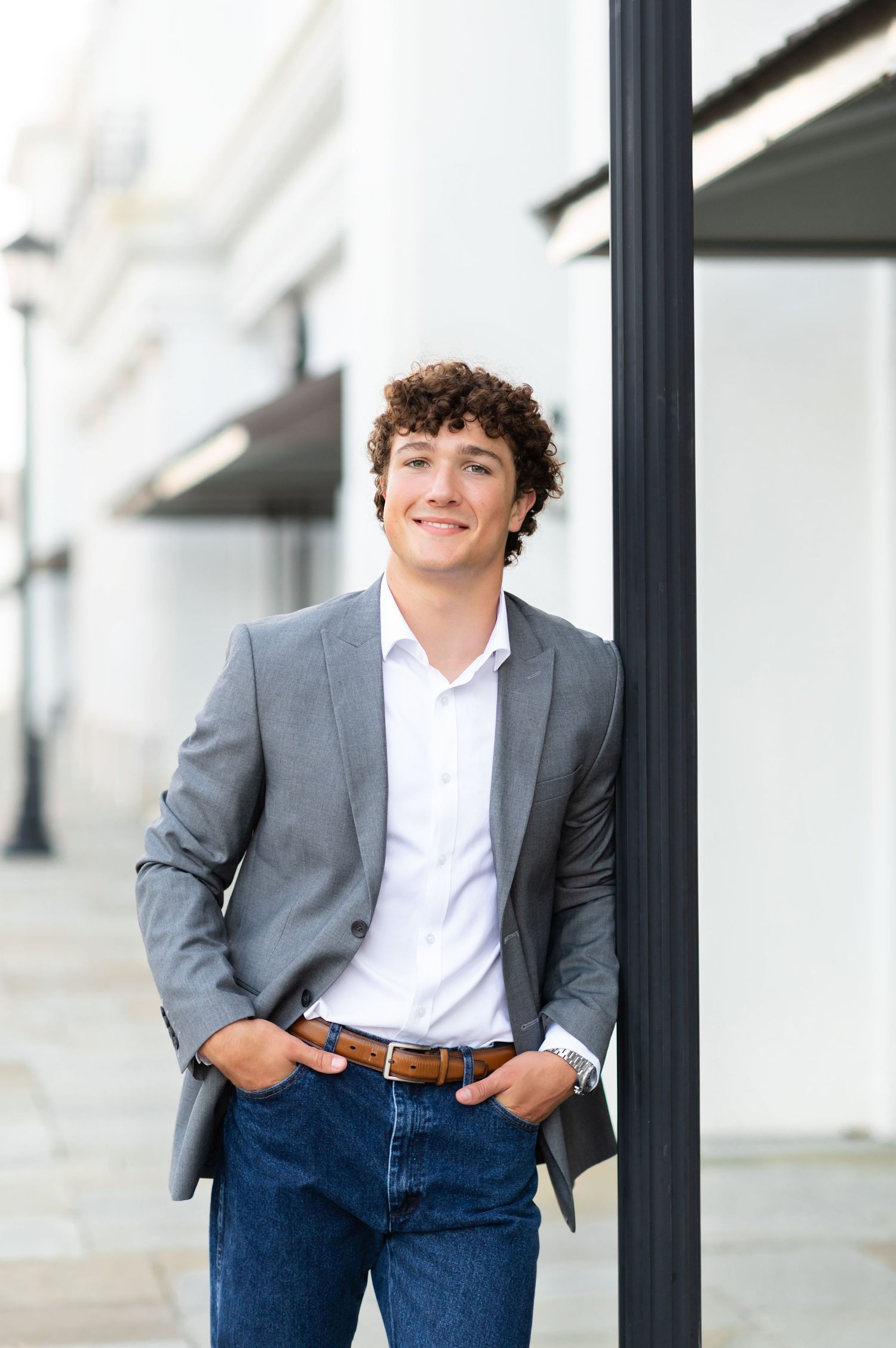 Young man with curly hair in gray blazer, white shirt, and jeans leaning against a black pole outdoors.