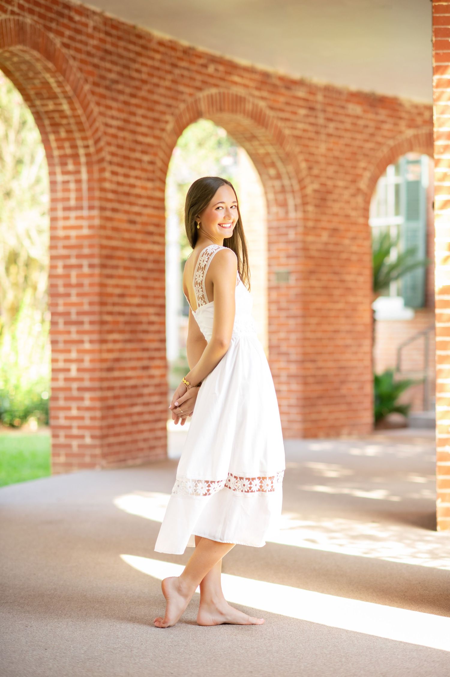 Young woman in white midi dress standing barefoot in sunlit brick archway corridor, smiling over shoulder.