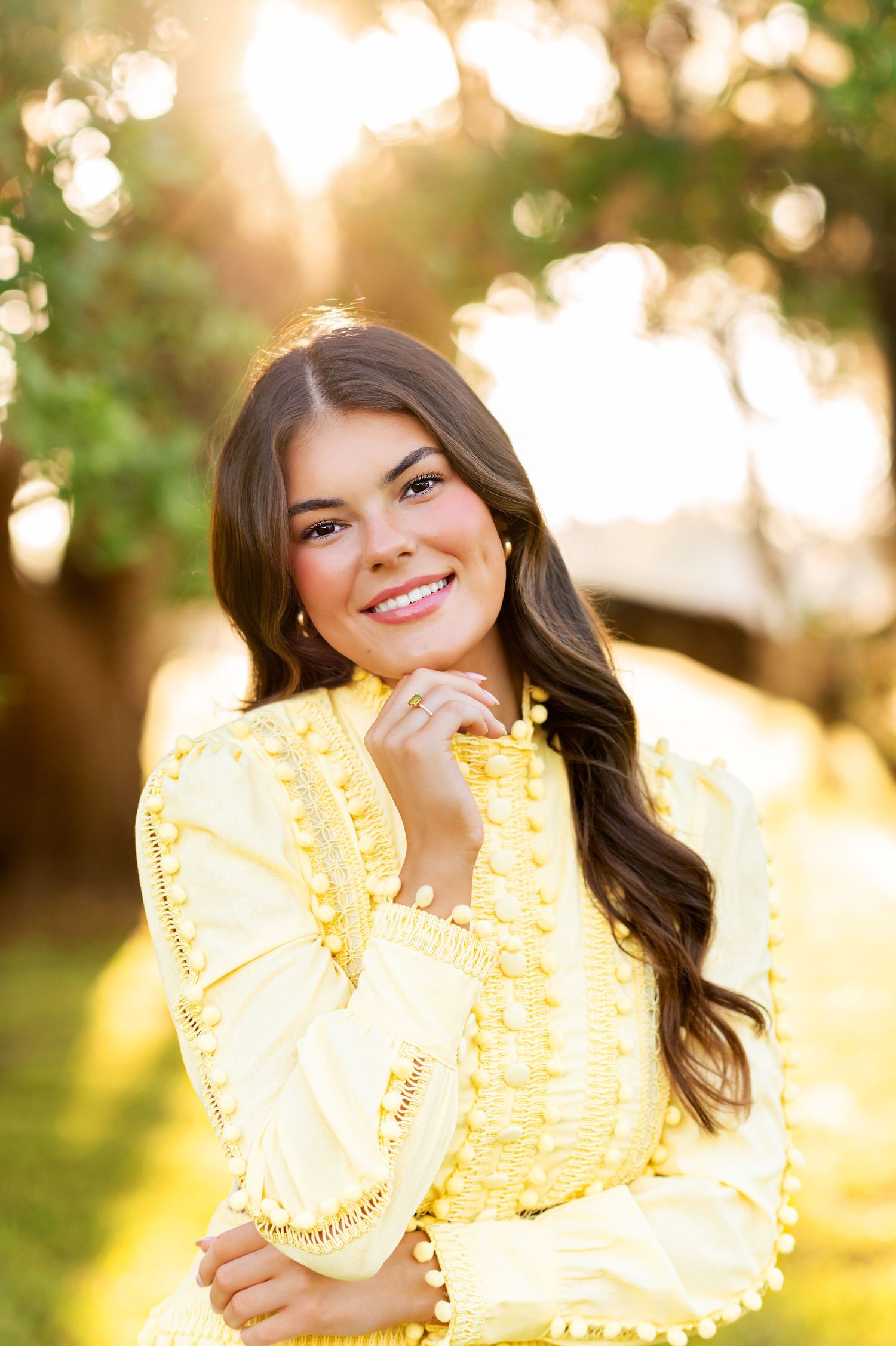 Young woman in yellow lace dress smiling outdoors with golden sunlight filtering through green trees behind her.
