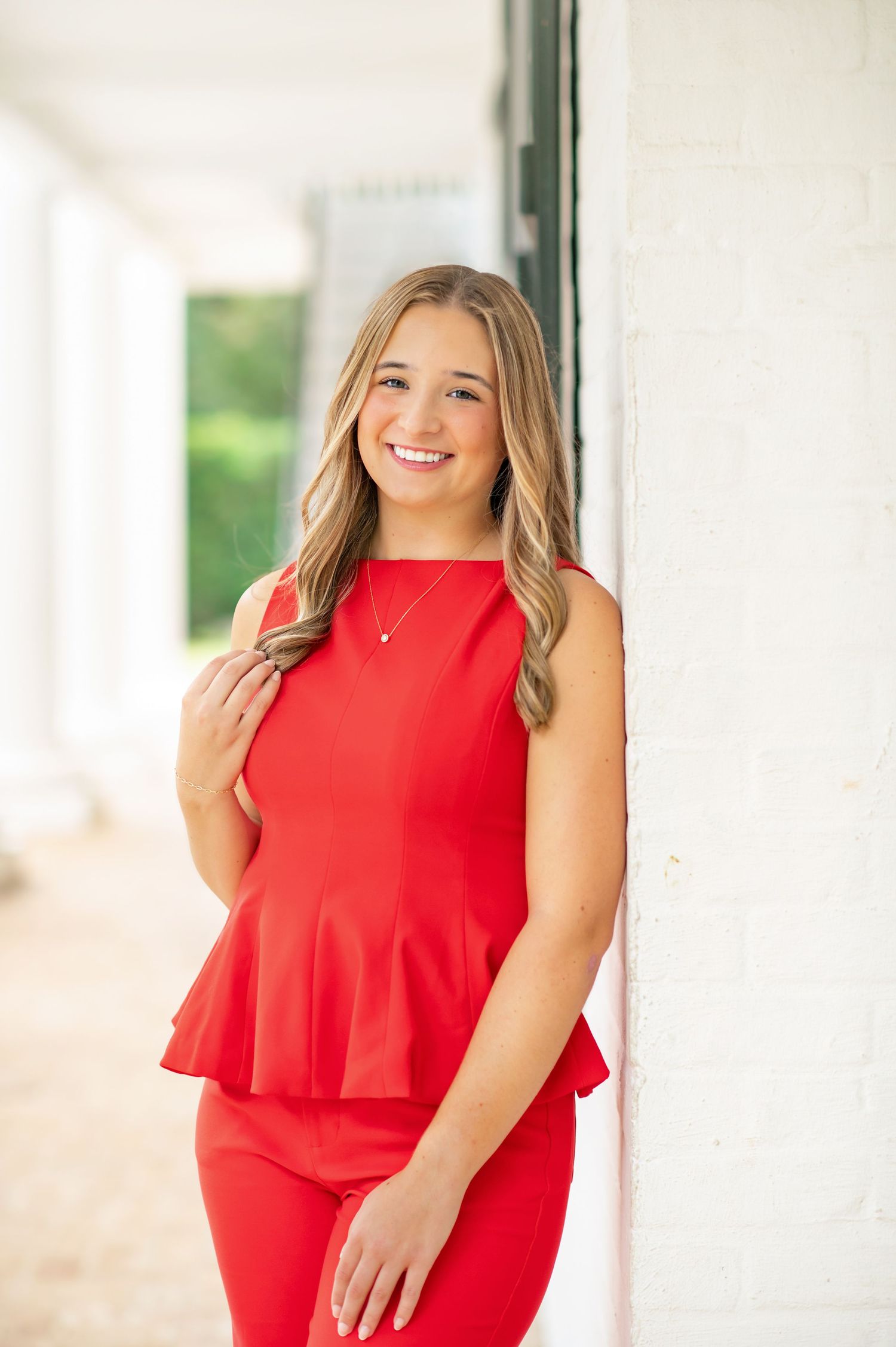 Young woman in red peplum outfit smiling and leaning against white wall in outdoor corridor.