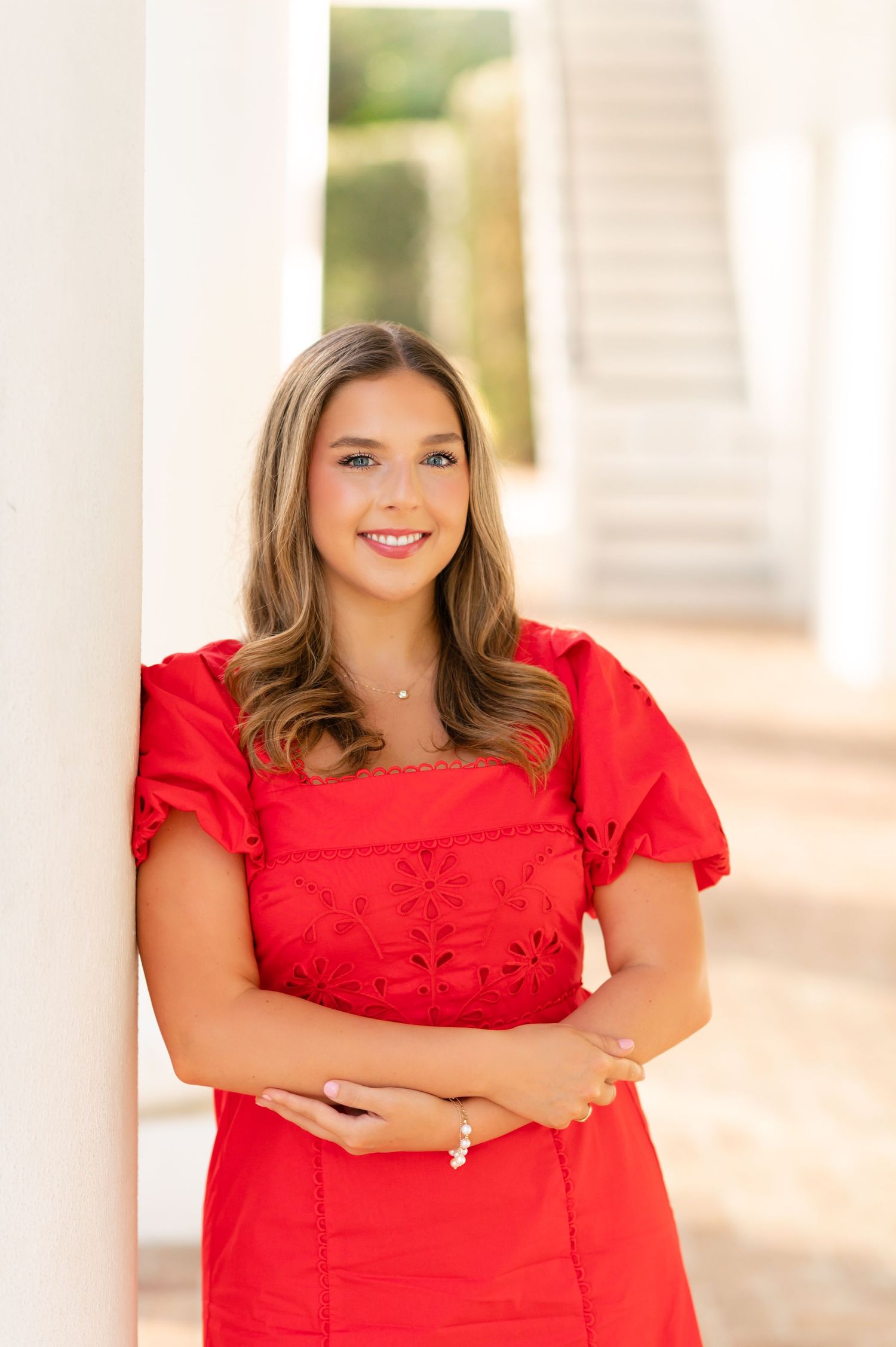 Smiling young woman in red embroidered dress leans against white column in elegant outdoor setting.