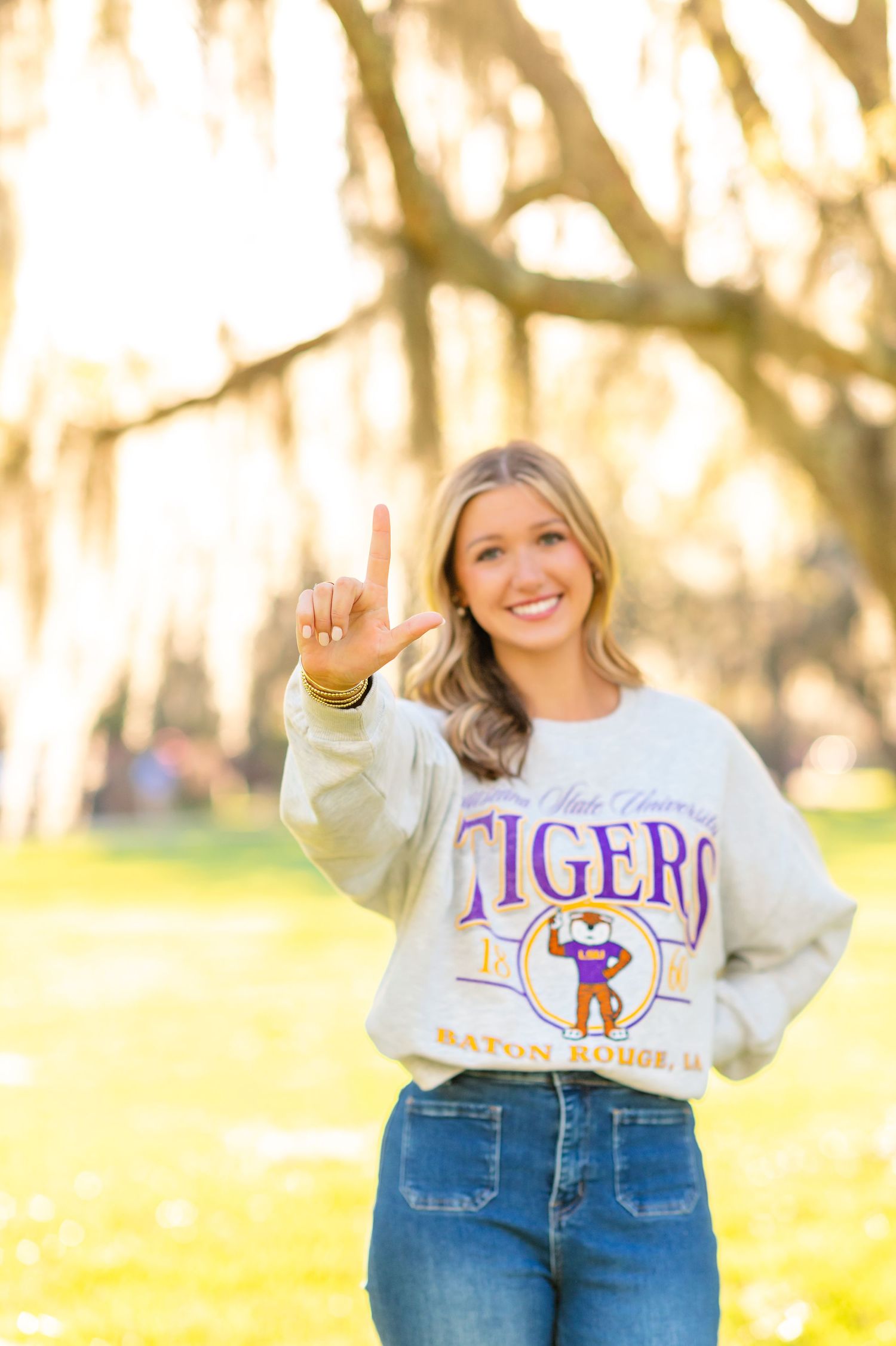 Smiling blonde woman wearing LSU Tigers Baton Rouge sweatshirt making number one hand gesture outdoors.