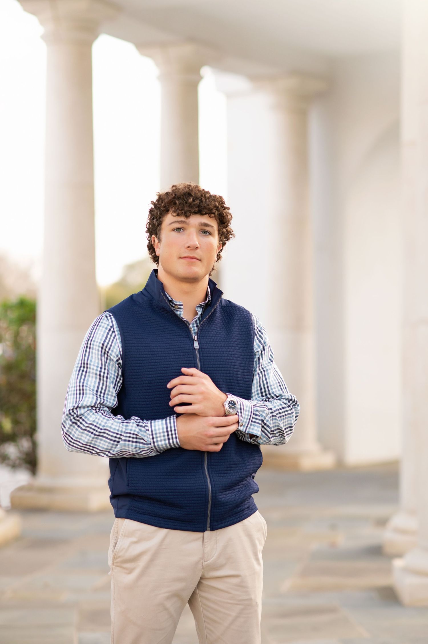 Young man with curly hair wearing a navy vest and plaid shirt poses confidently near white columns.