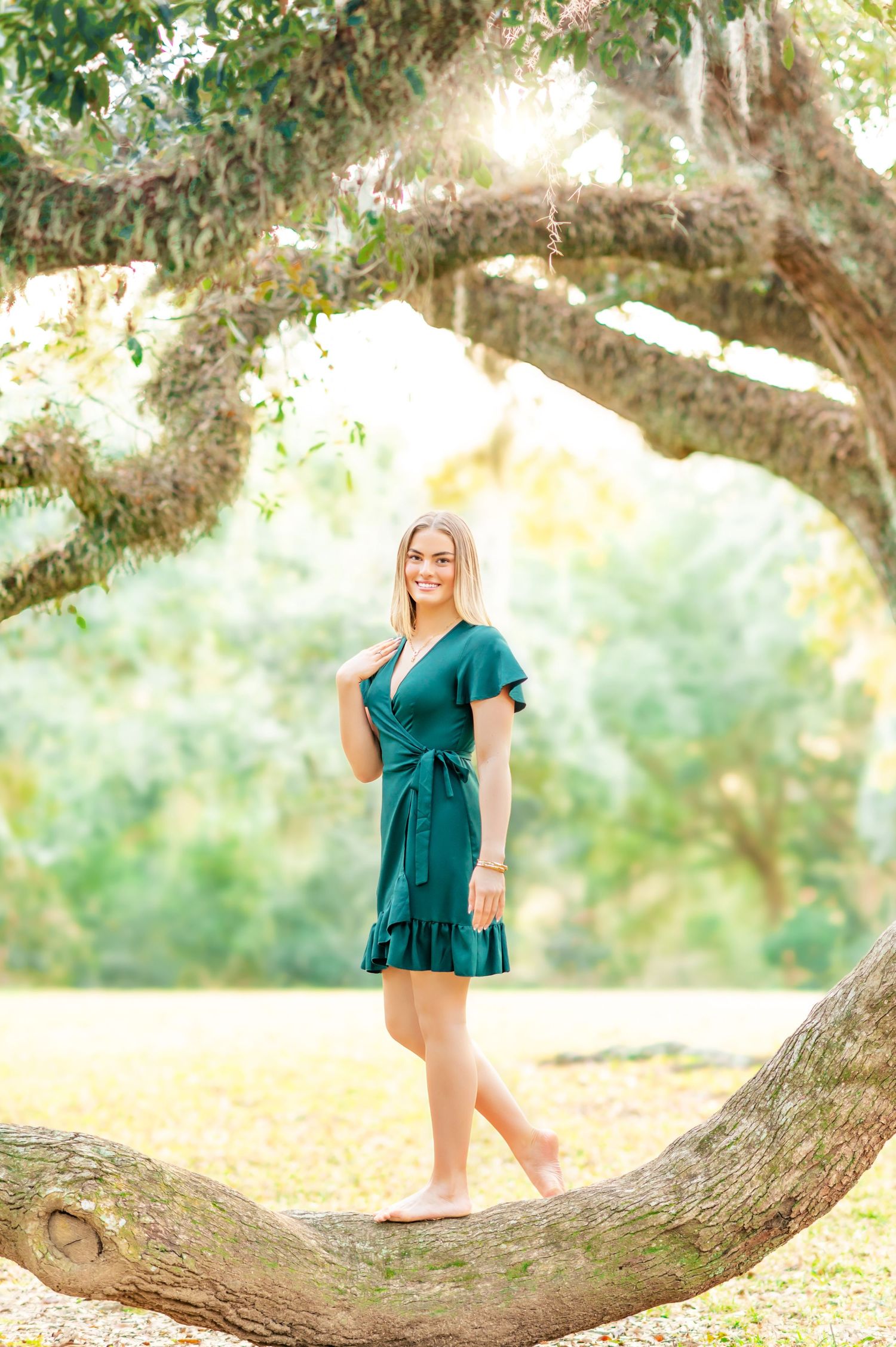 Blonde teen girl in emerald wrap dress balances barefoot on large oak tree branch in sunny park.