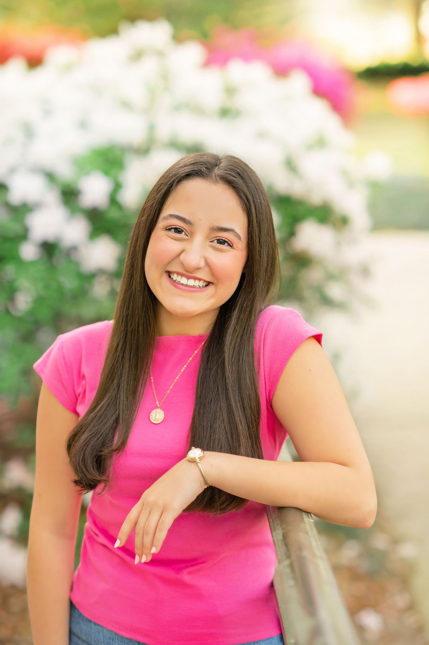 Smiling brunette woman in hot pink t-shirt leans on a railing with colorful flowers in background.