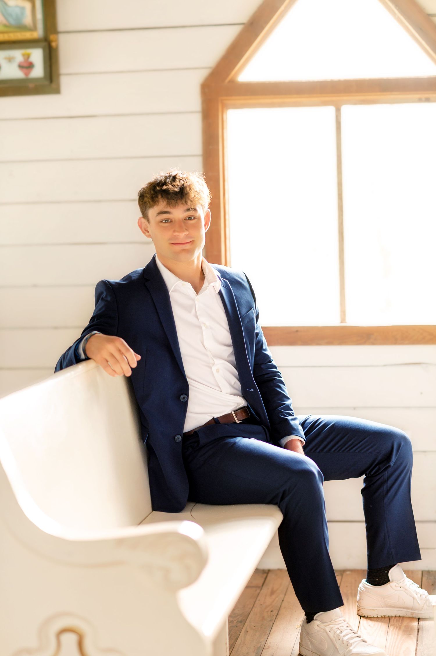 Young man in navy blue suit sitting on white chair near bright window in white shiplap room.