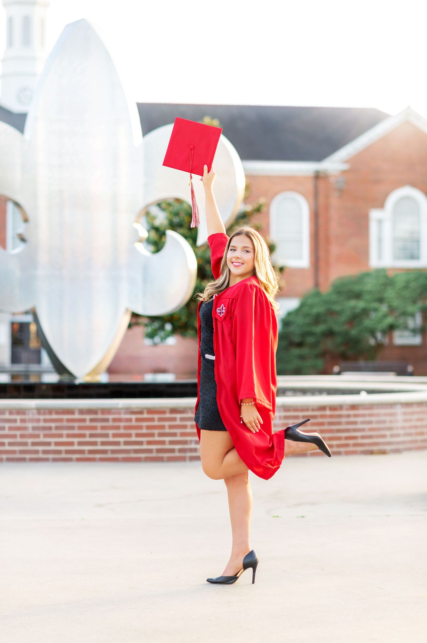 Joyful graduate celebrates in red cap and gown, raising mortarboard outside university building.