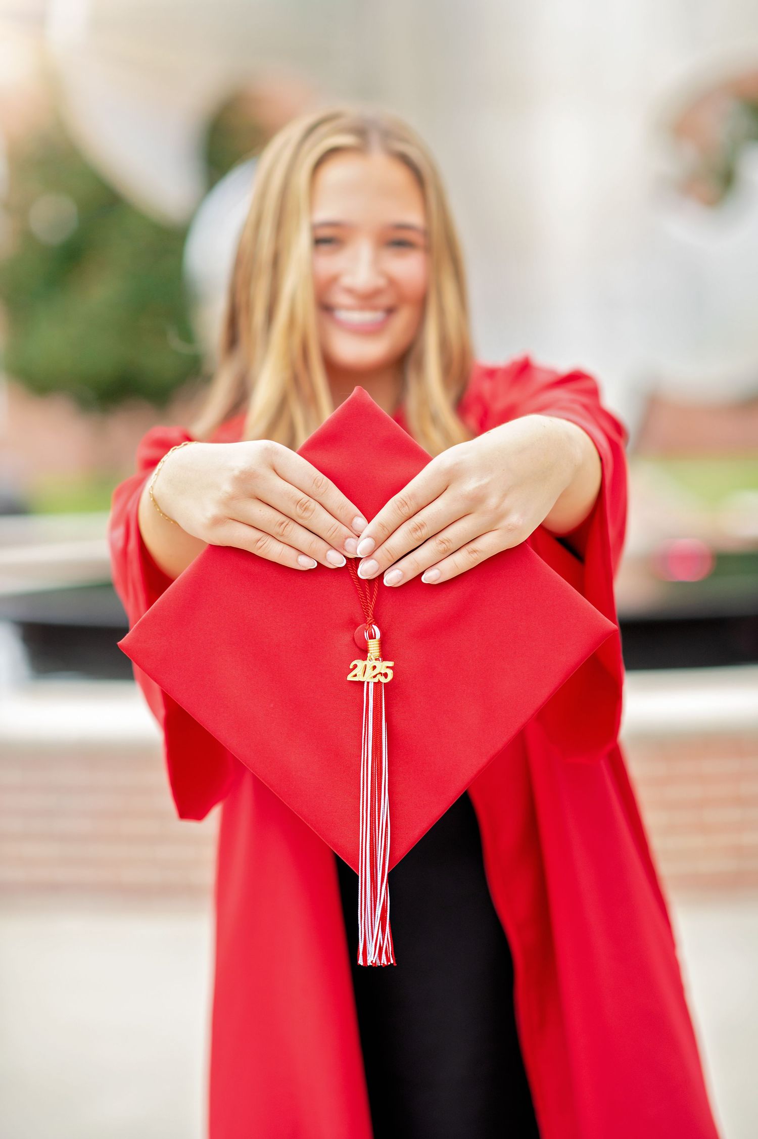 Smiling graduate in red cap and gown holding her 2025 graduation cap toward camera outdoors.
