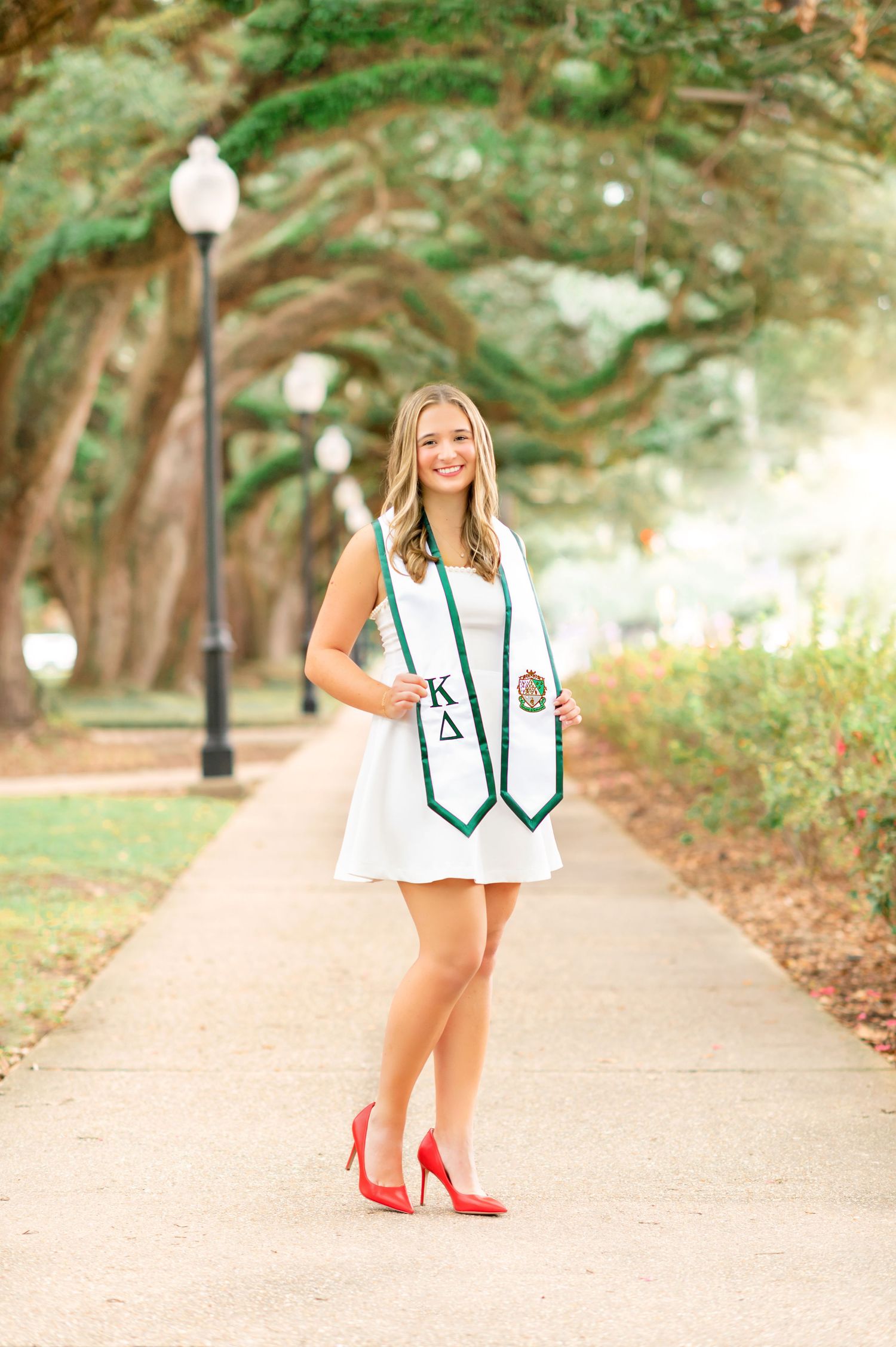 Smiling graduate in white dress and green Greek letter stole poses on tree-lined campus path wearing red heels.