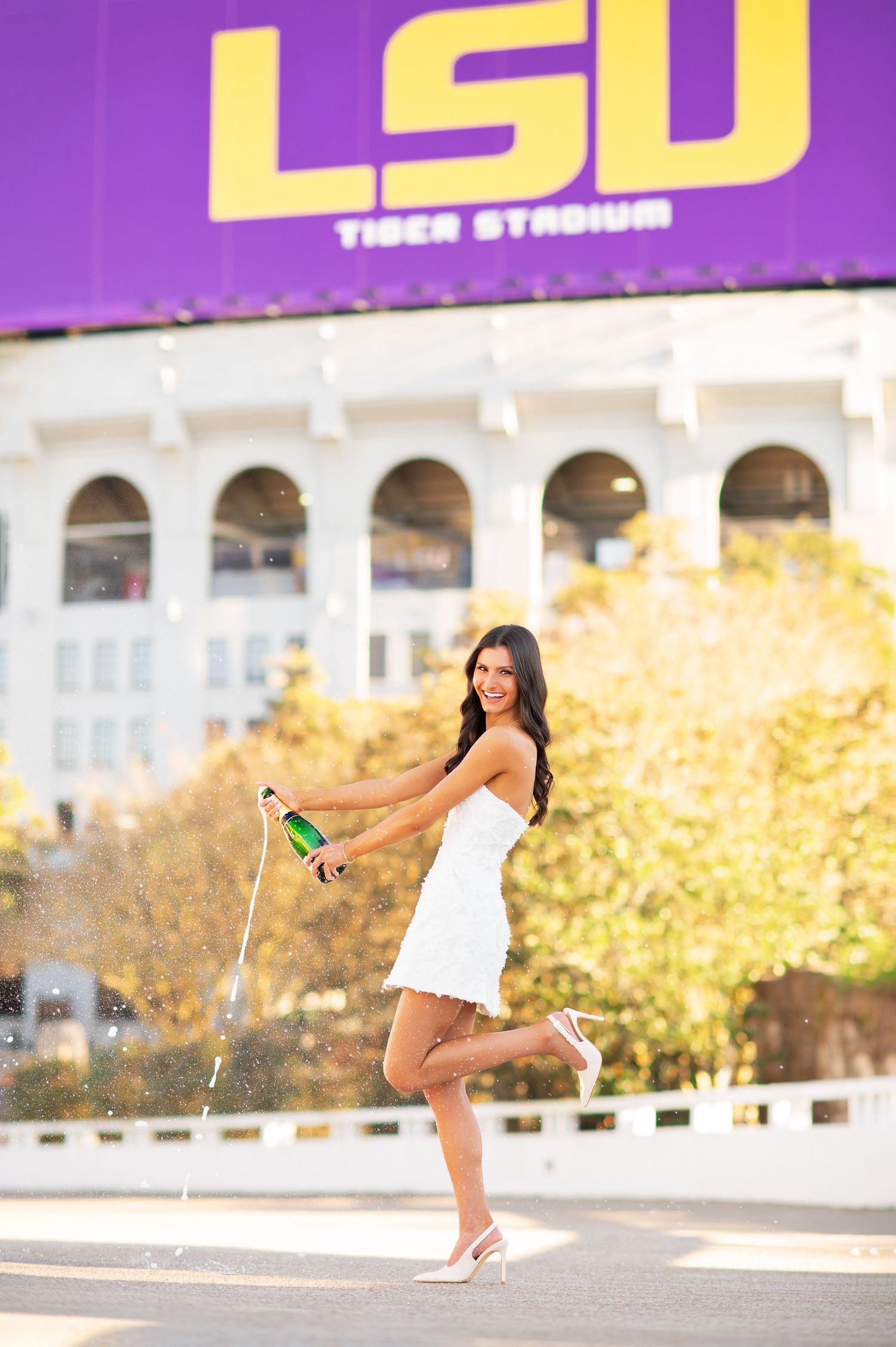 A woman in a white dress pops champagne in front of LSU Tiger Stadium celebrating her graduation.