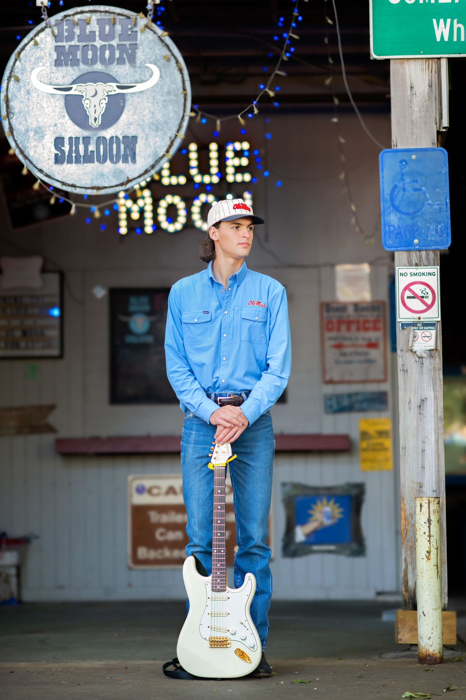 Young musician stands with white Fender Stratocaster guitar outside Blue Moon Saloon wearing denim shirt and trucker hat.