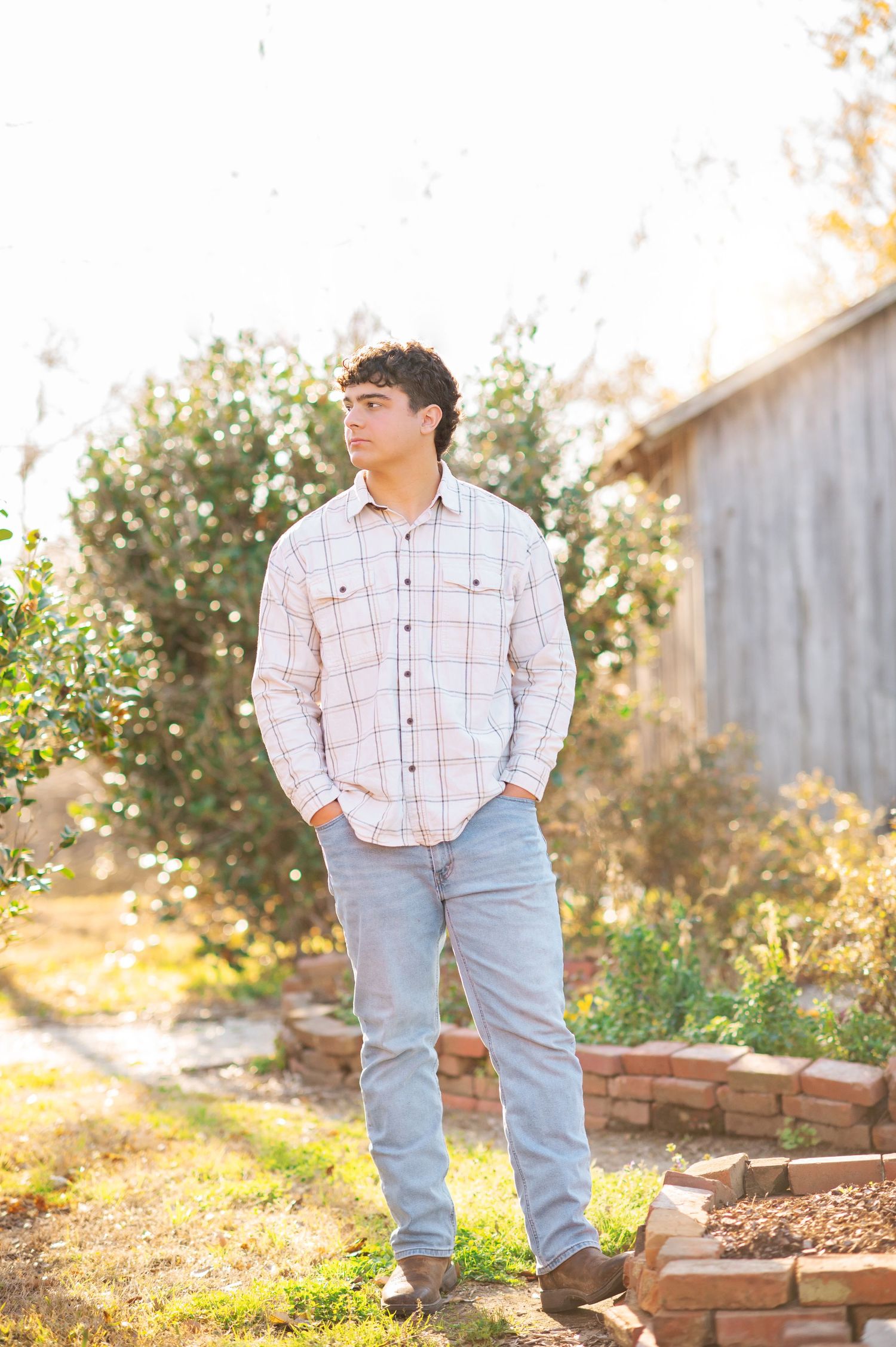 Young man in plaid shirt and jeans standing outdoors near brick garden border and wooden fence on sunny day.