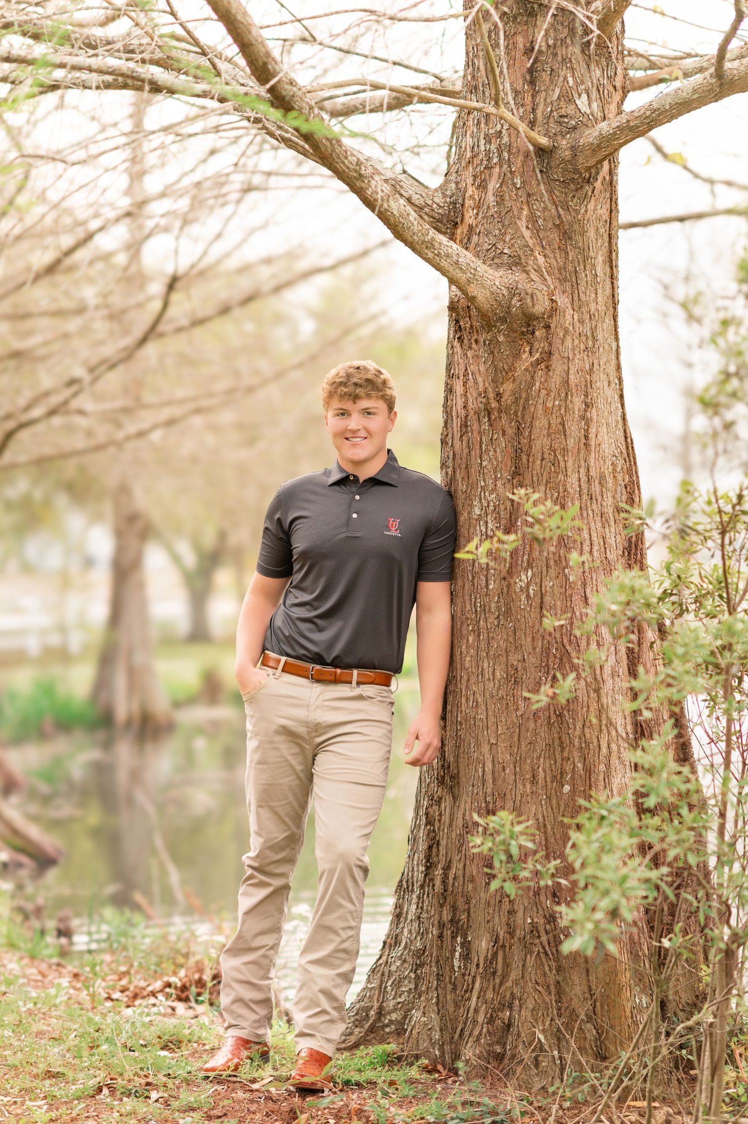 Smiling young man in dark polo shirt leaning against a tree near a pond outdoors.