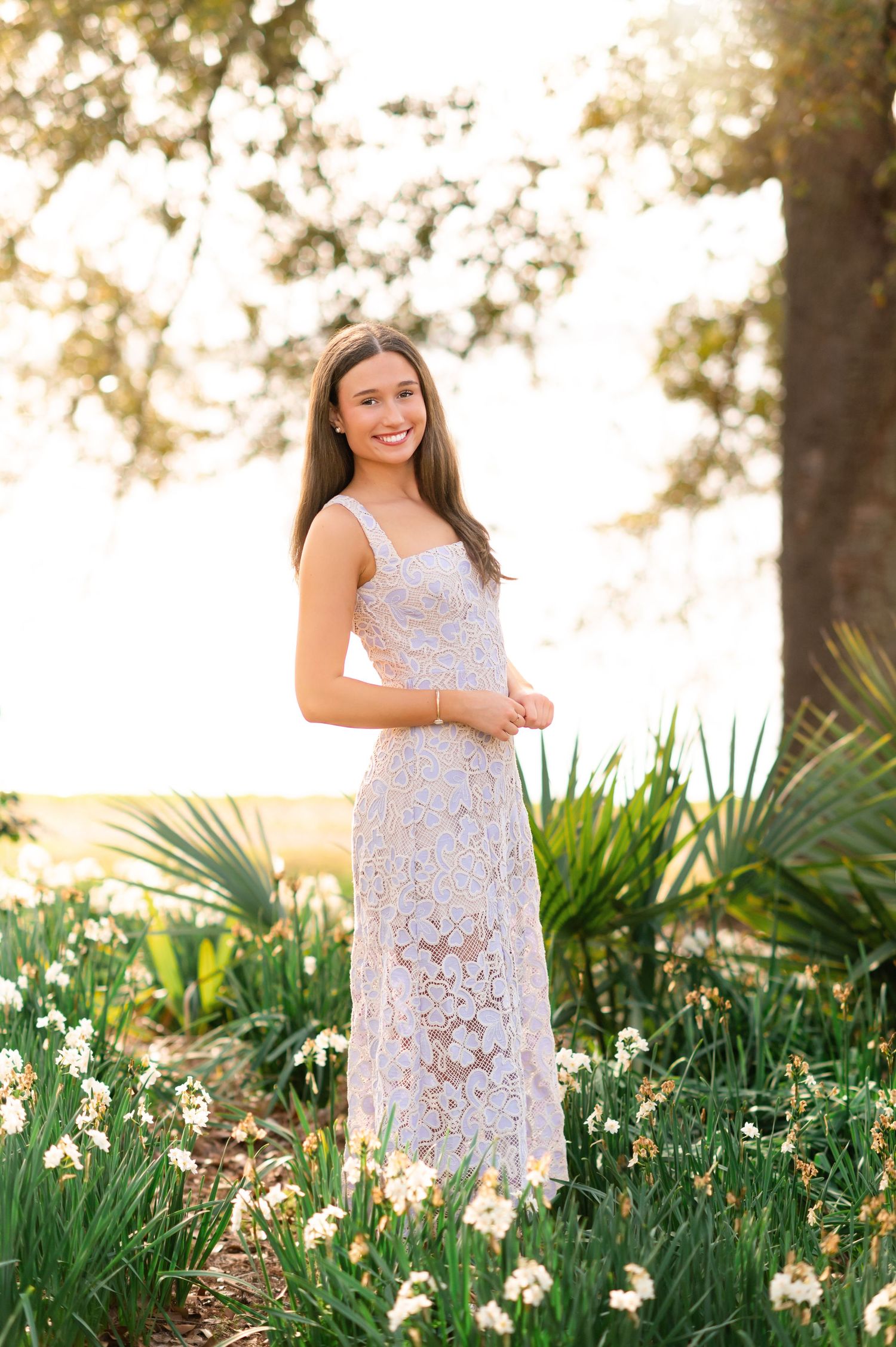 Young woman in white lace maxi dress smiling outdoors among daffodils and tropical plants in golden sunlight.