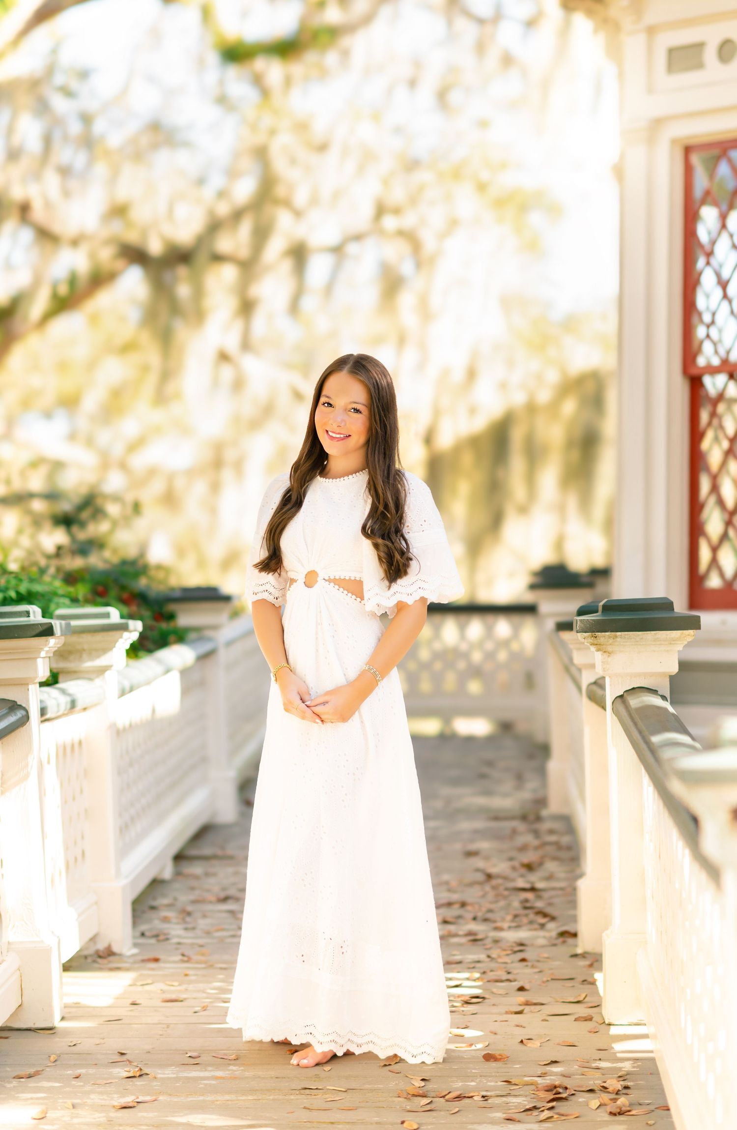 Young woman in white lace maxi dress poses on sunlit Southern plantation porch with Spanish moss trees.