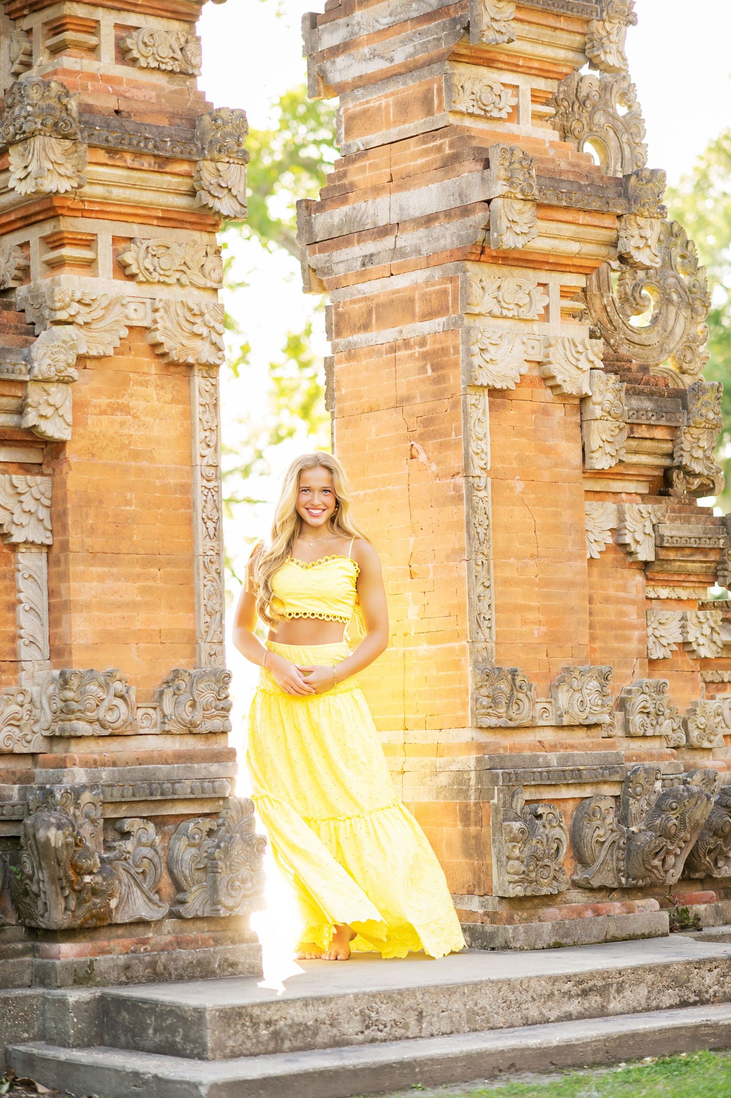Blonde woman in yellow two-piece outfit posing between ornate stone temple columns outdoors.