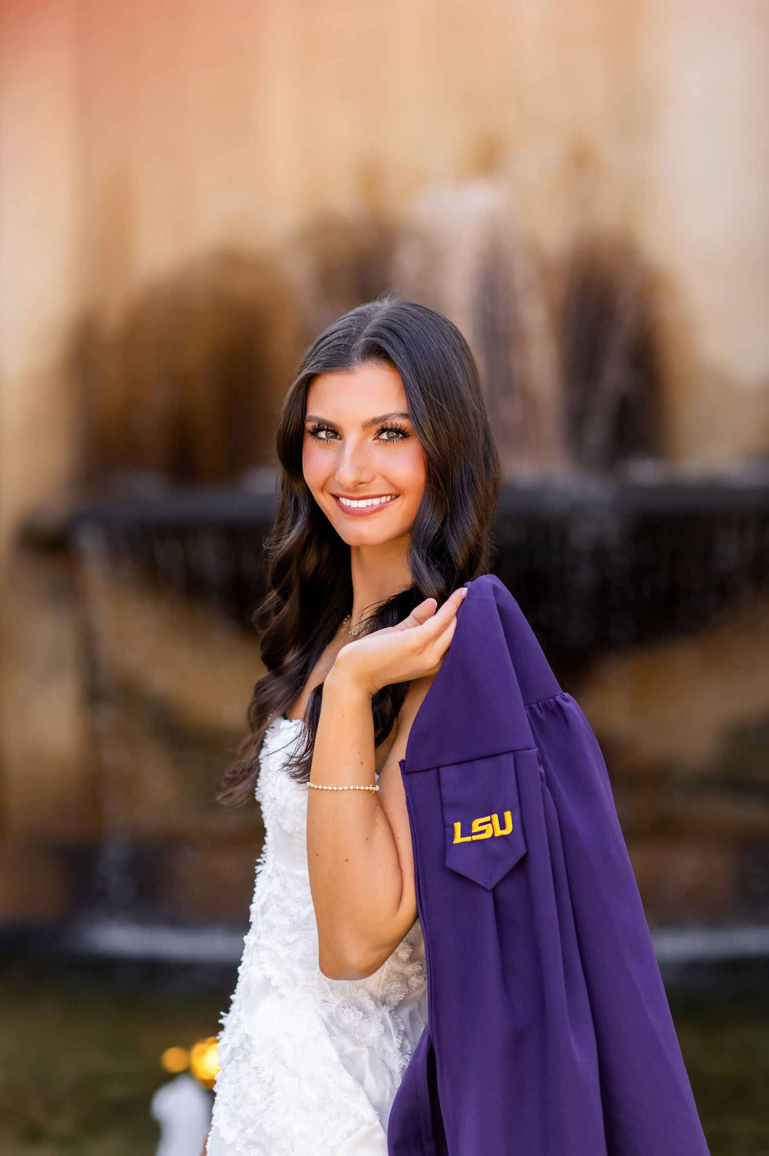 Smiling LSU graduate poses with purple graduation gown over shoulder in front of fountain.