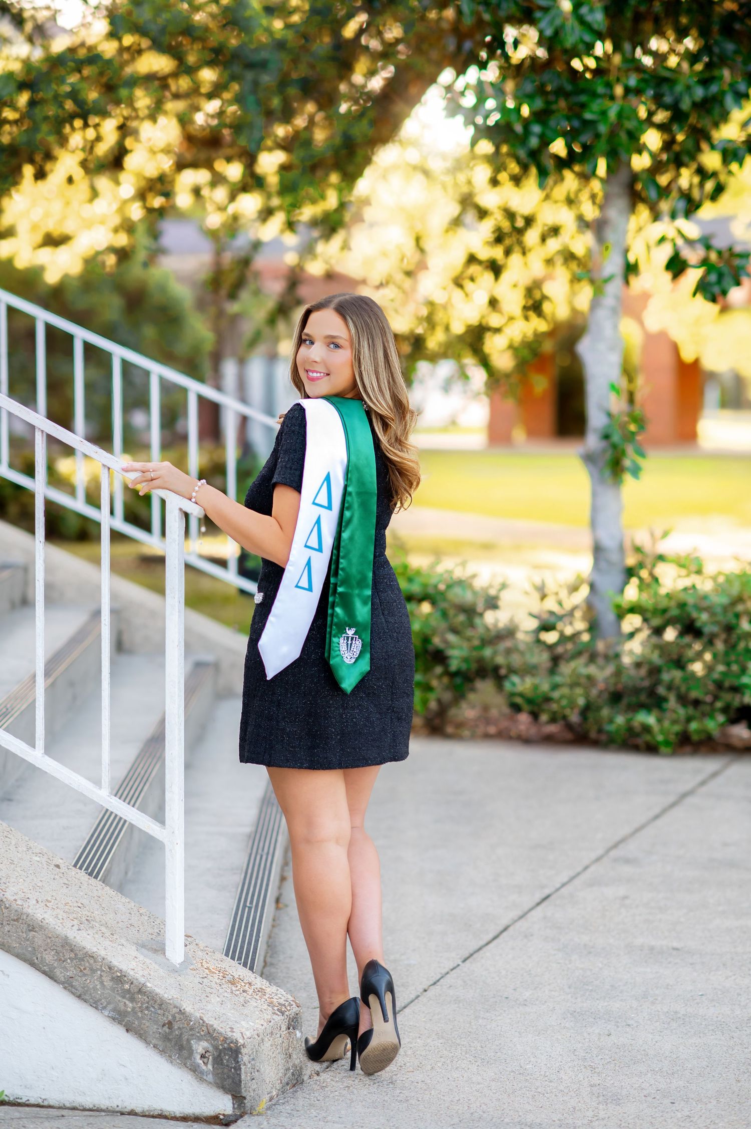 Young woman in black dress with Delta Delta Delta graduation stoles poses on sunlit outdoor staircase.