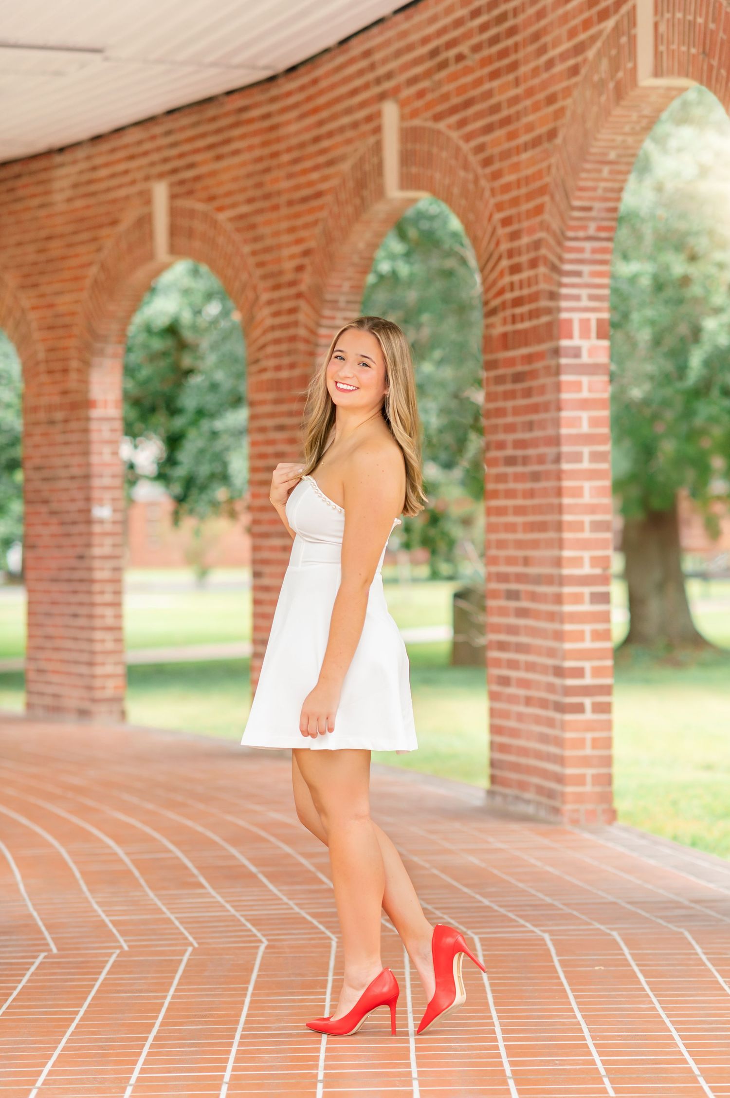 Young woman in white dress and red heels posing under brick arches on a sunny day.