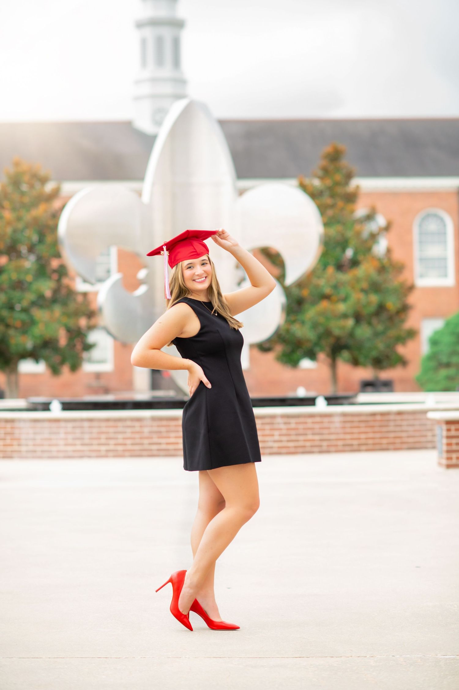 Young woman in black dress and red heels poses outdoors in graduation cap, smiling confidently on campus.