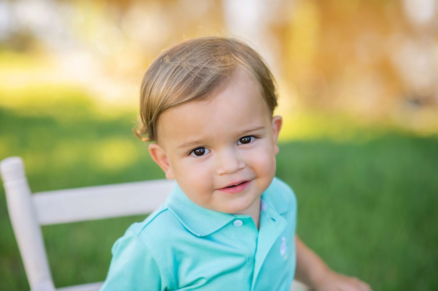 Smiling toddler boy in teal polo shirt sitting on white chair outdoors on green grass.