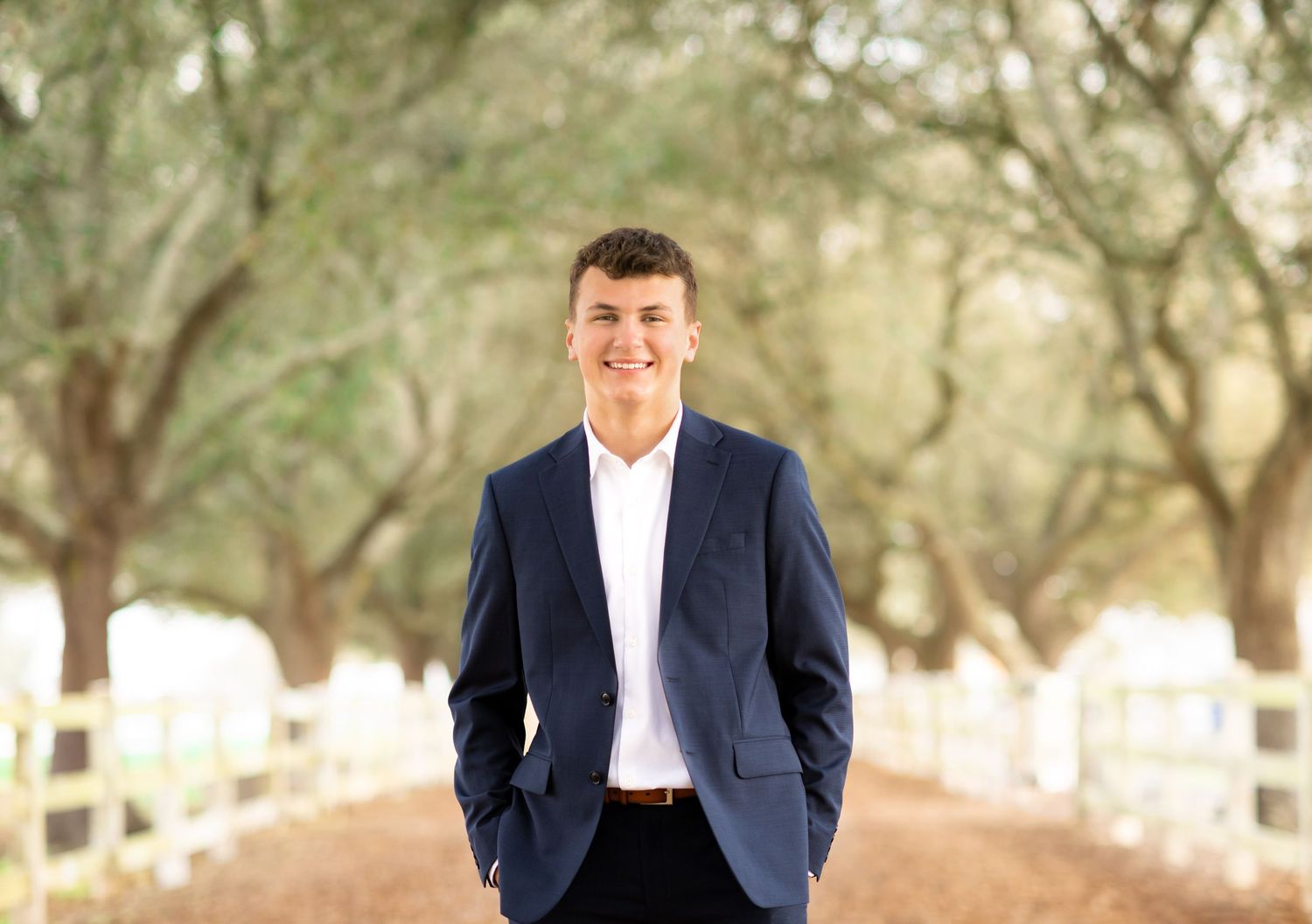 Young man in navy blazer smiling on a tree-lined dirt path with white fences, ideal senior portrait photo.