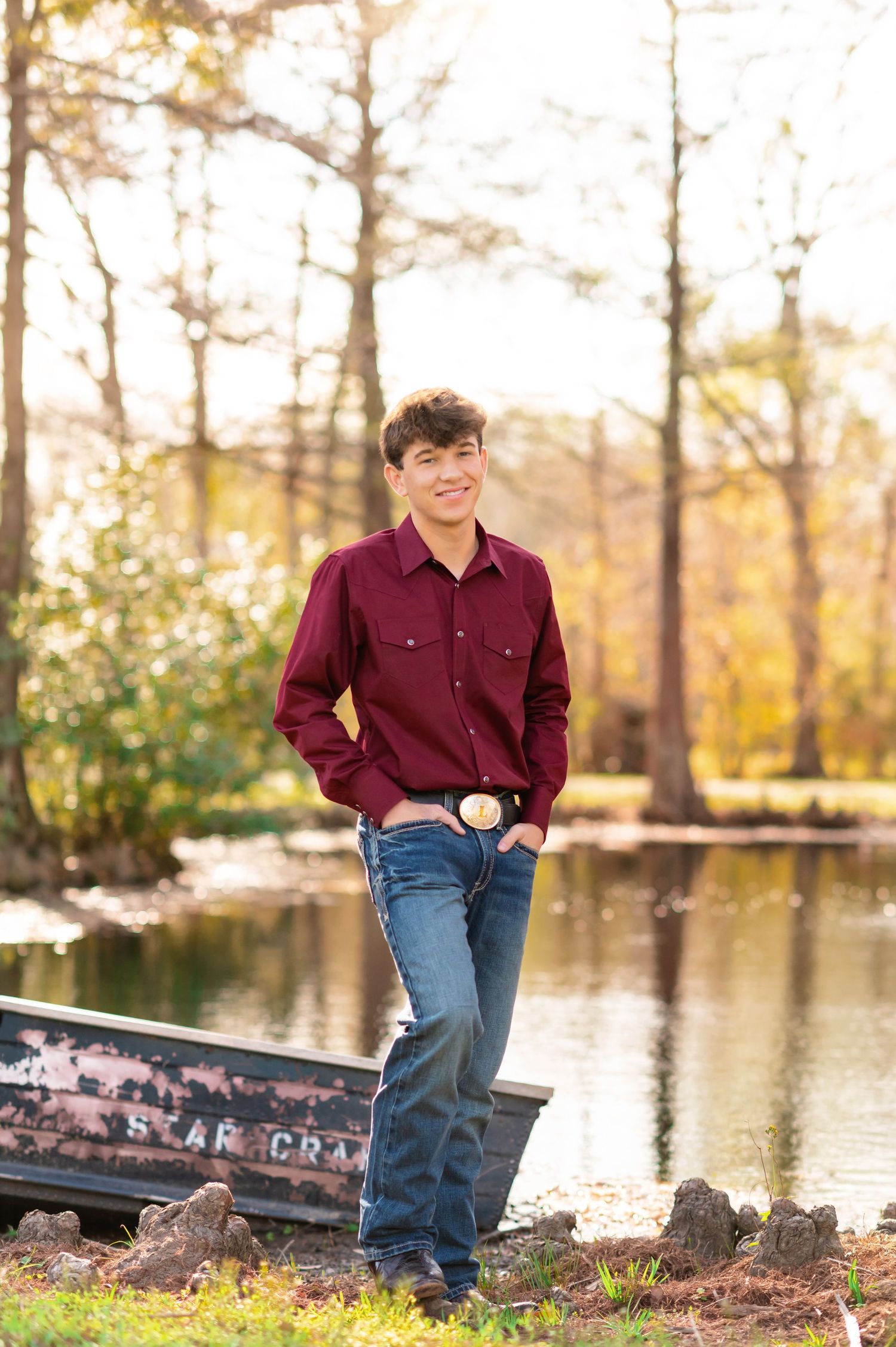 Young man in maroon shirt and jeans smiling by a pond with autumn trees in background.
