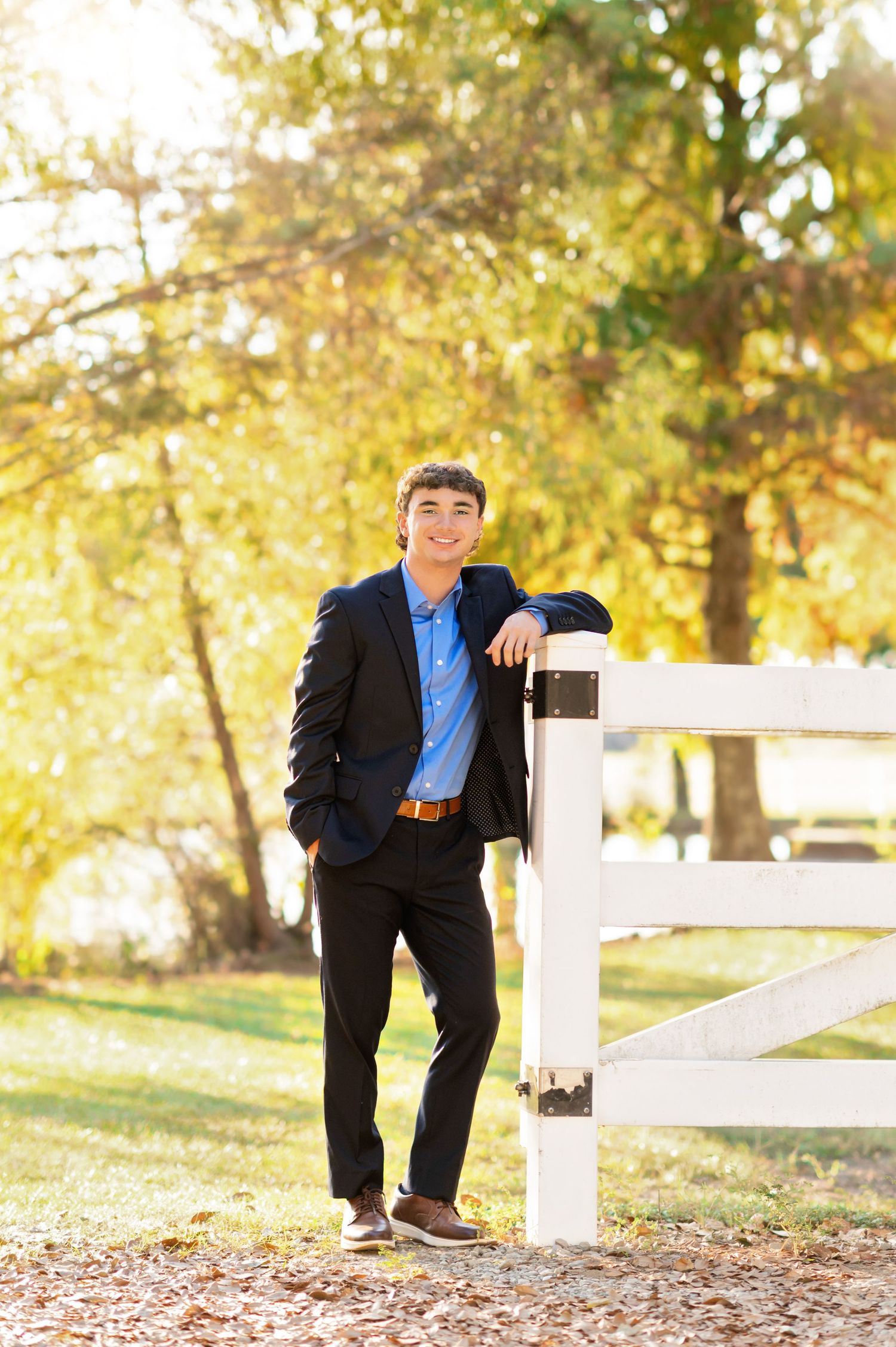 Young man in blue shirt and blazer leaning on white fence outdoors during golden autumn afternoon.