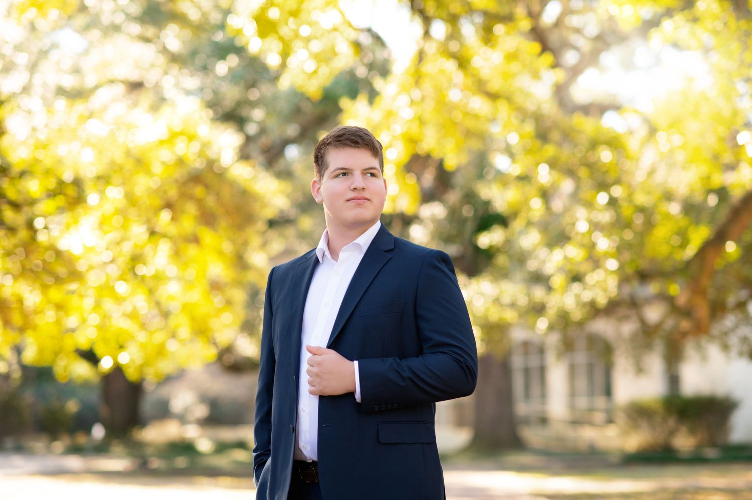 Professional man in navy blazer poses outdoors among golden autumn trees in a polished portrait photo.