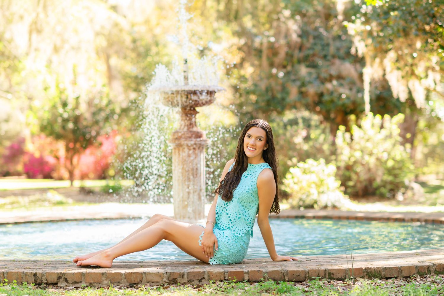 Young woman in teal lace outfit sitting by a decorative fountain in a lush green park on a sunny day.
