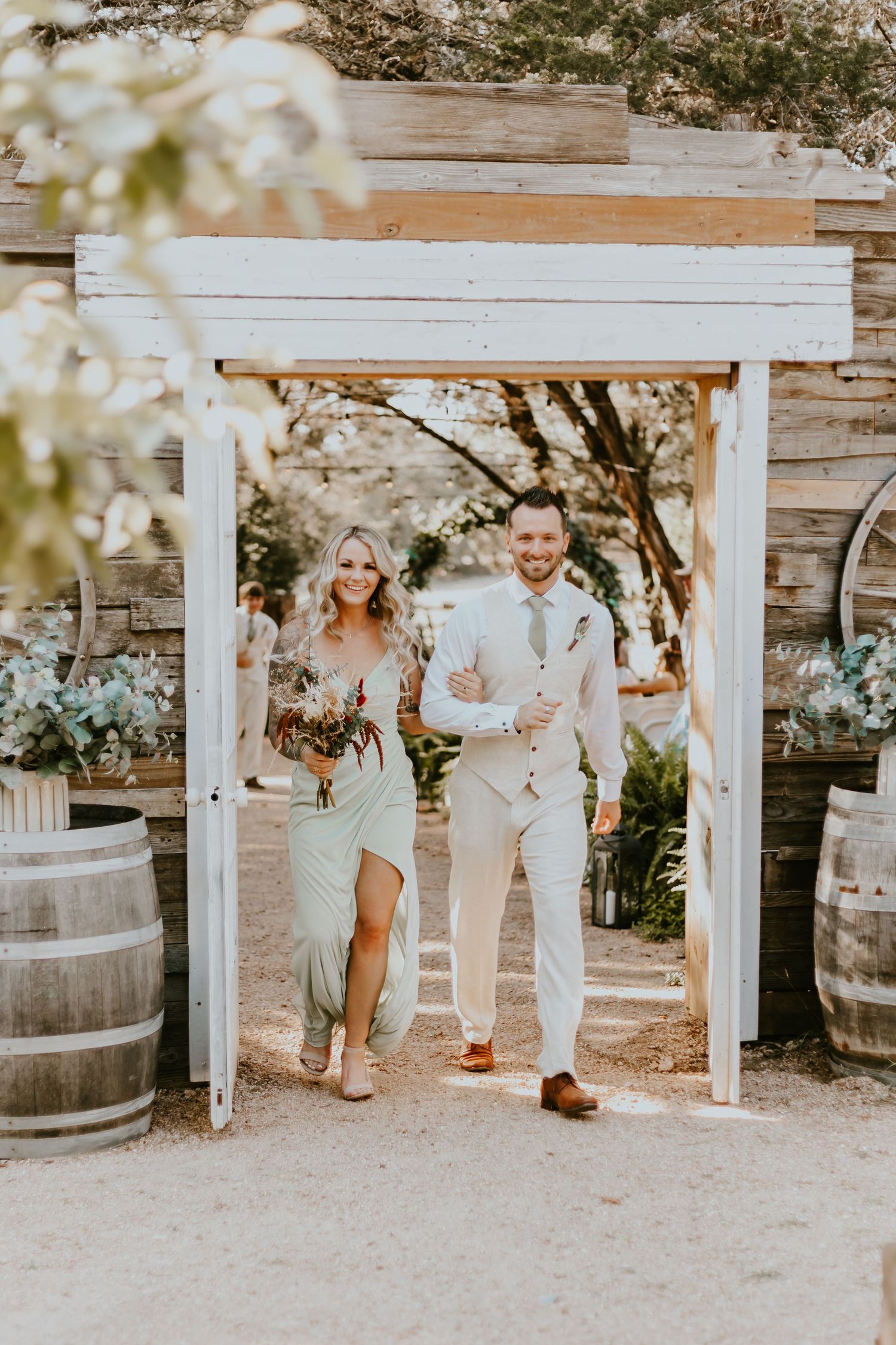 A couple walks through a rustic white wooden archway decorated with wine barrels at an outdoor venue.