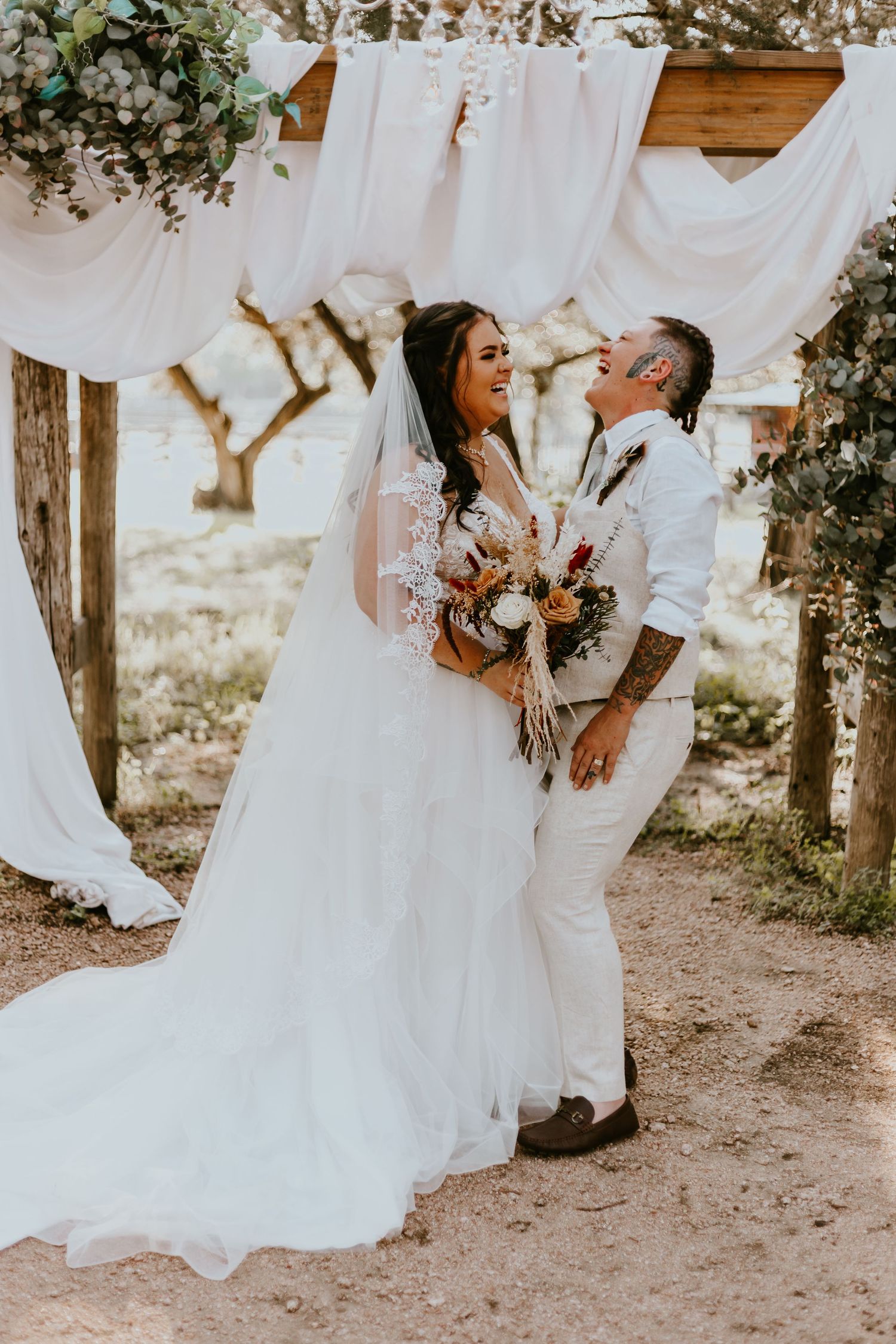 A newlywed couple shares a romantic moment under flowing white fabric and floral decorations at an outdoor rustic wedding.