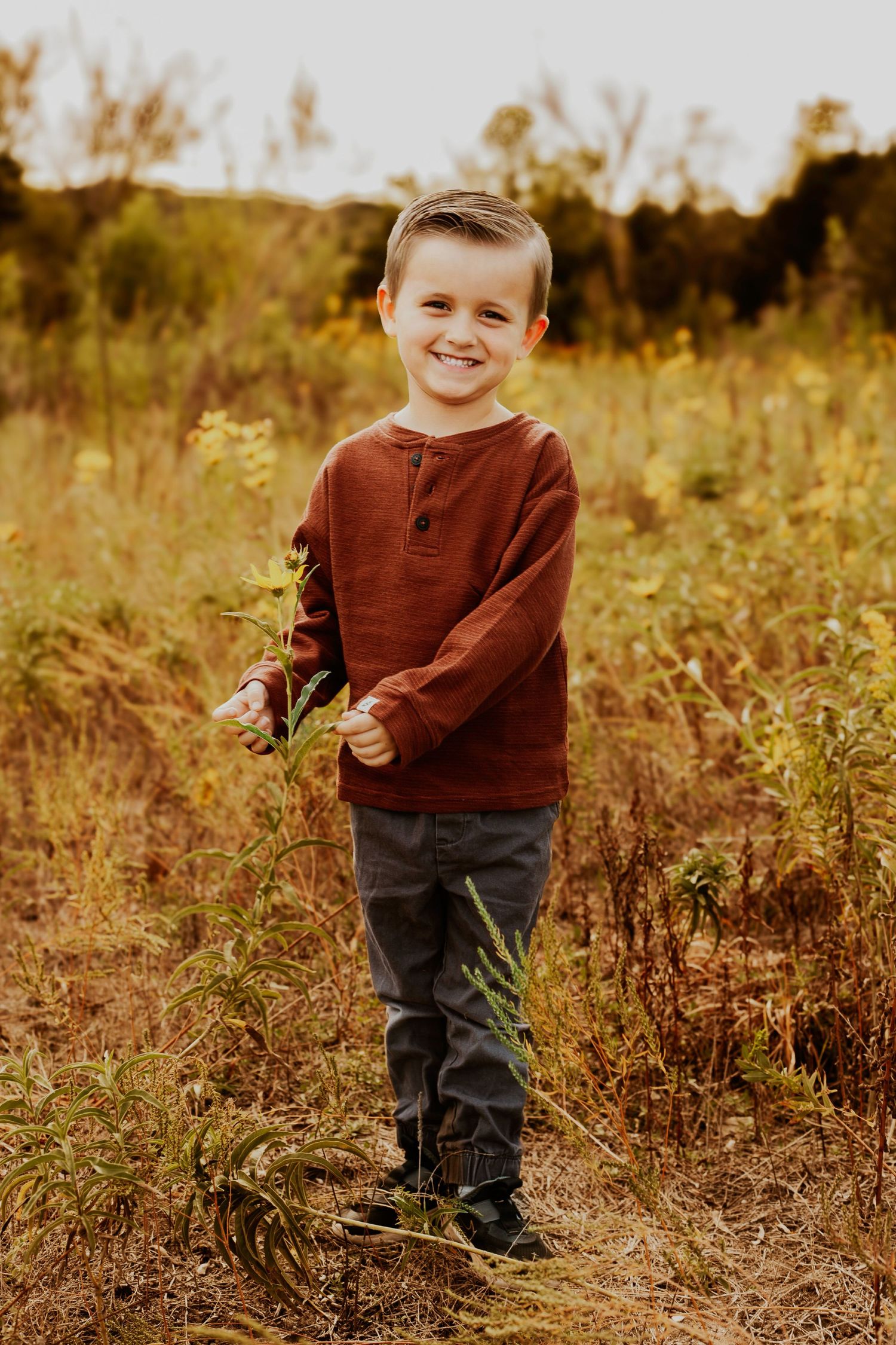 Young child stands in golden autumn field wearing rust-colored sweater and dark pants.