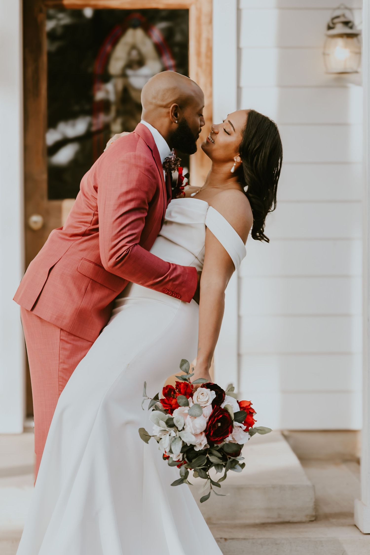 Romantic wedding couple embraces in a tender dip kiss, bride holding red and white bouquet against white backdrop.