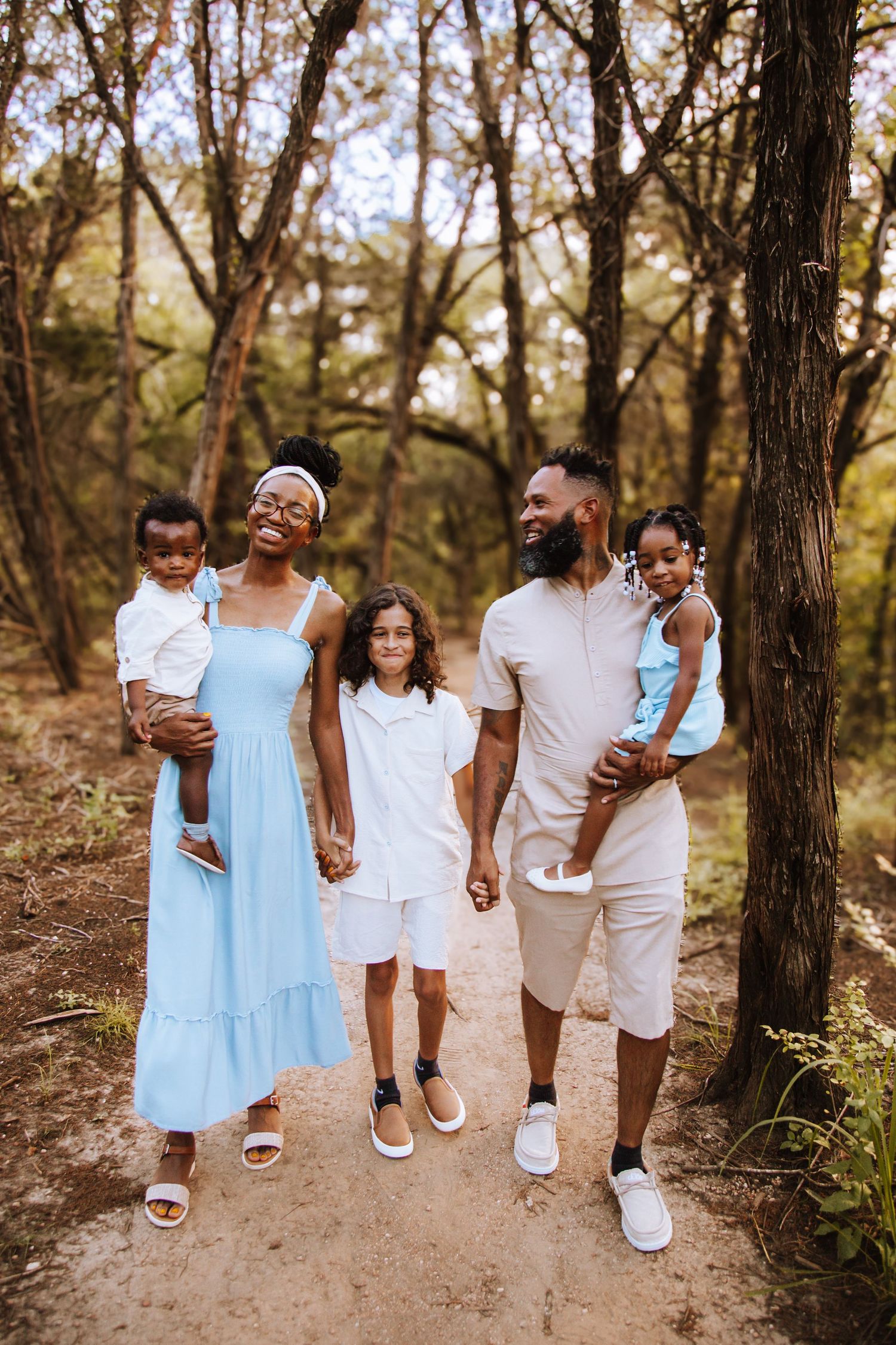A family in coordinated white and light blue outfits stands together on a wooded trail during a sunny day.