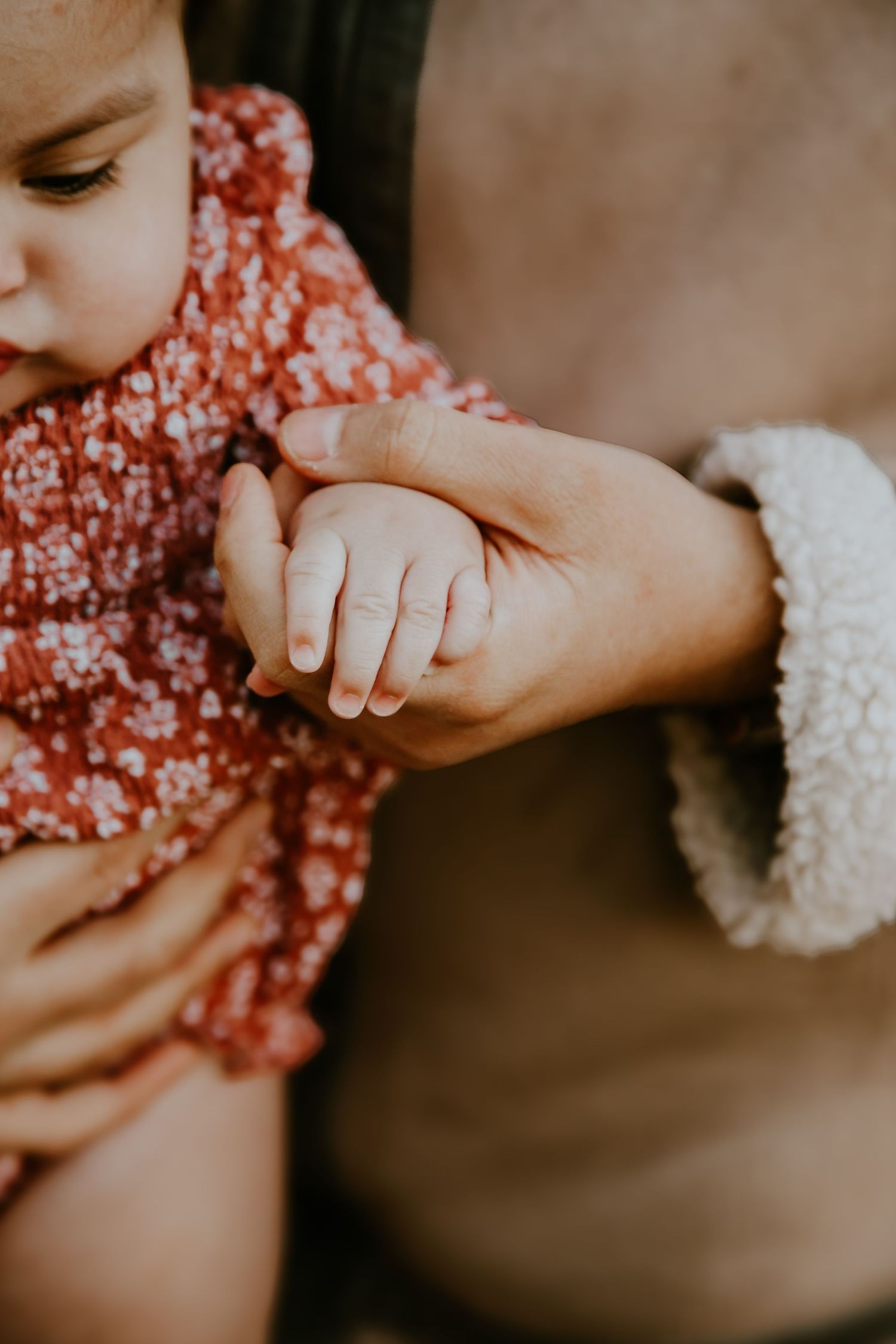 Close-up view of small baby hand being gently held by adult wearing white wool sweater and red floral dress.