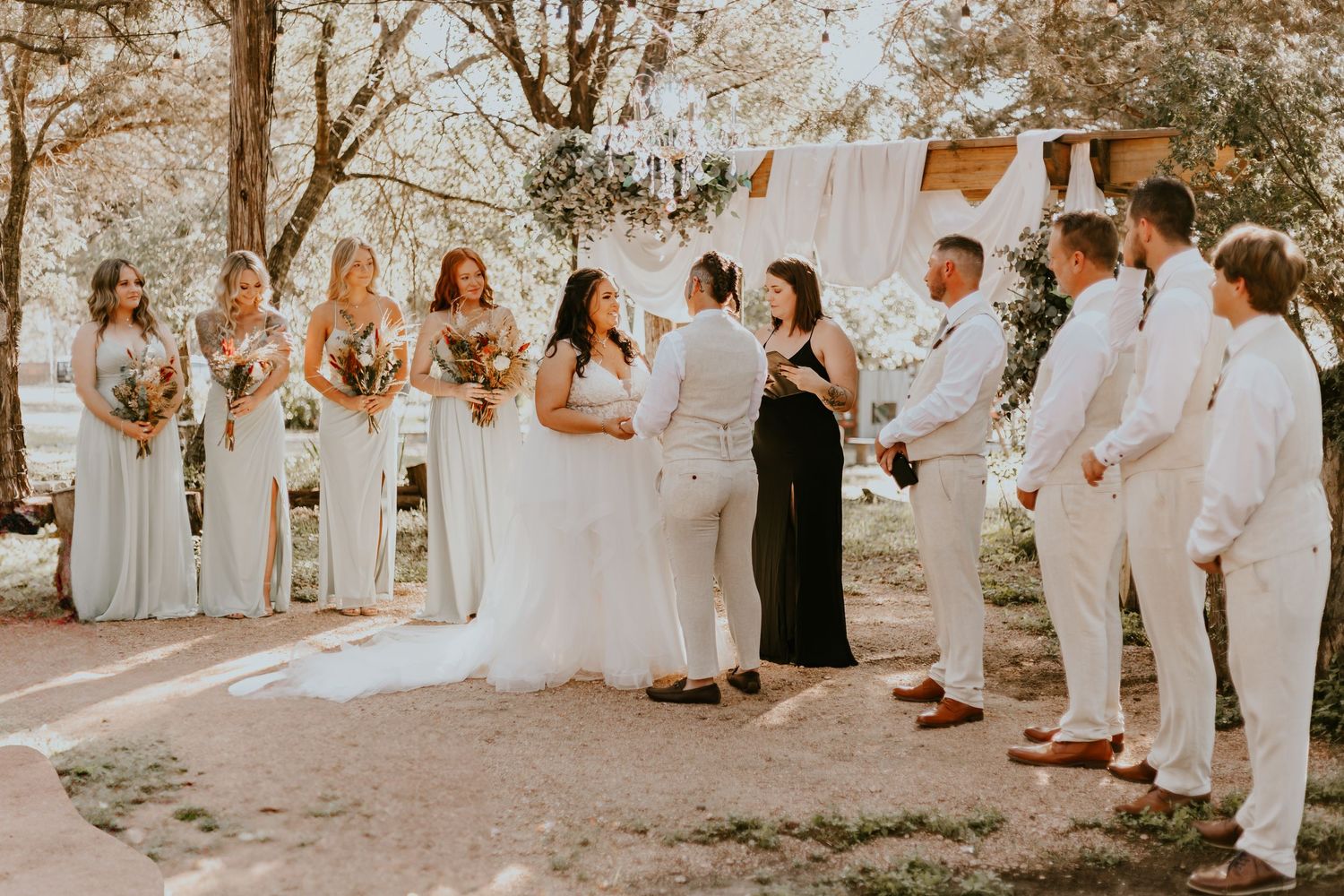 Wedding ceremony group gathers outdoors with bridesmaids in gray dresses and groomsmen wearing crisp white suits.